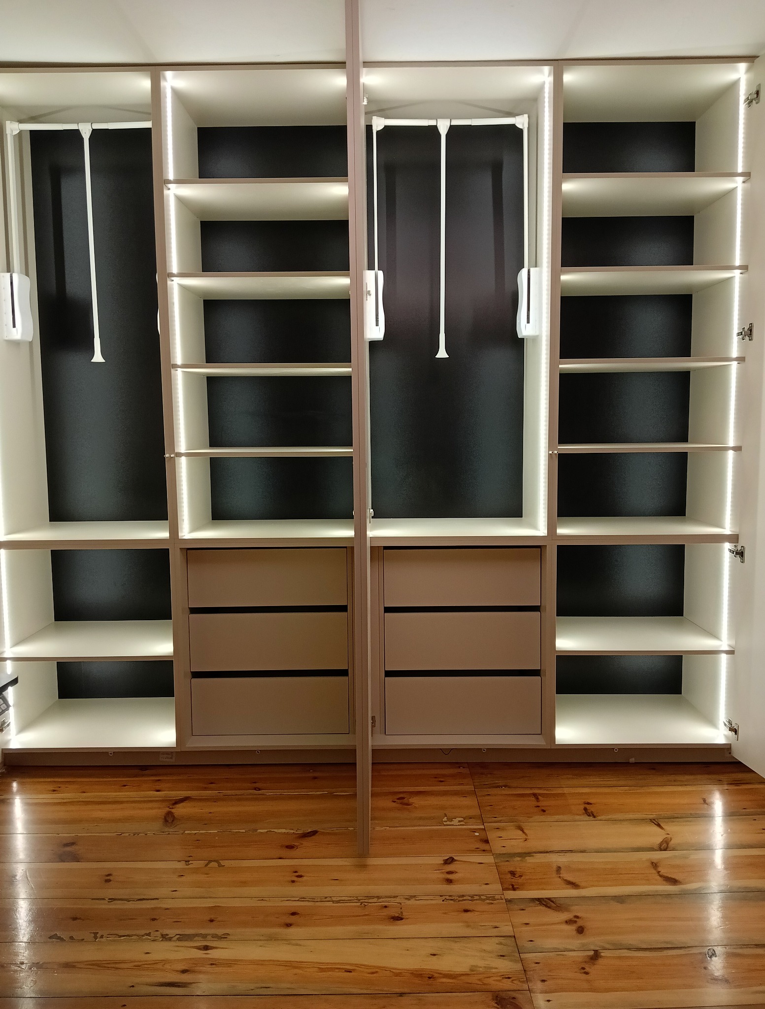 Otwarta szafa wnękowa z oświetleniem LED, widoczne półki, szuflady i drążki na ubrania. Na podłodze parkiet z naturalnego drewna. Minimalistyczny design.
