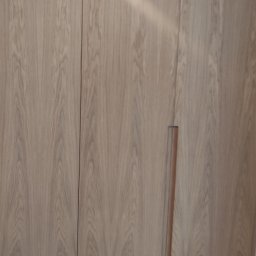 DEKOR MEBEL DESIGN Radosław Modzelewski - Jasna, drewniana szafa wnękowa z minimalistycznymi uchwytami, oświetlona górnym światłem. Widoczne subtelne usłojenie drewna i gładkie fronty.