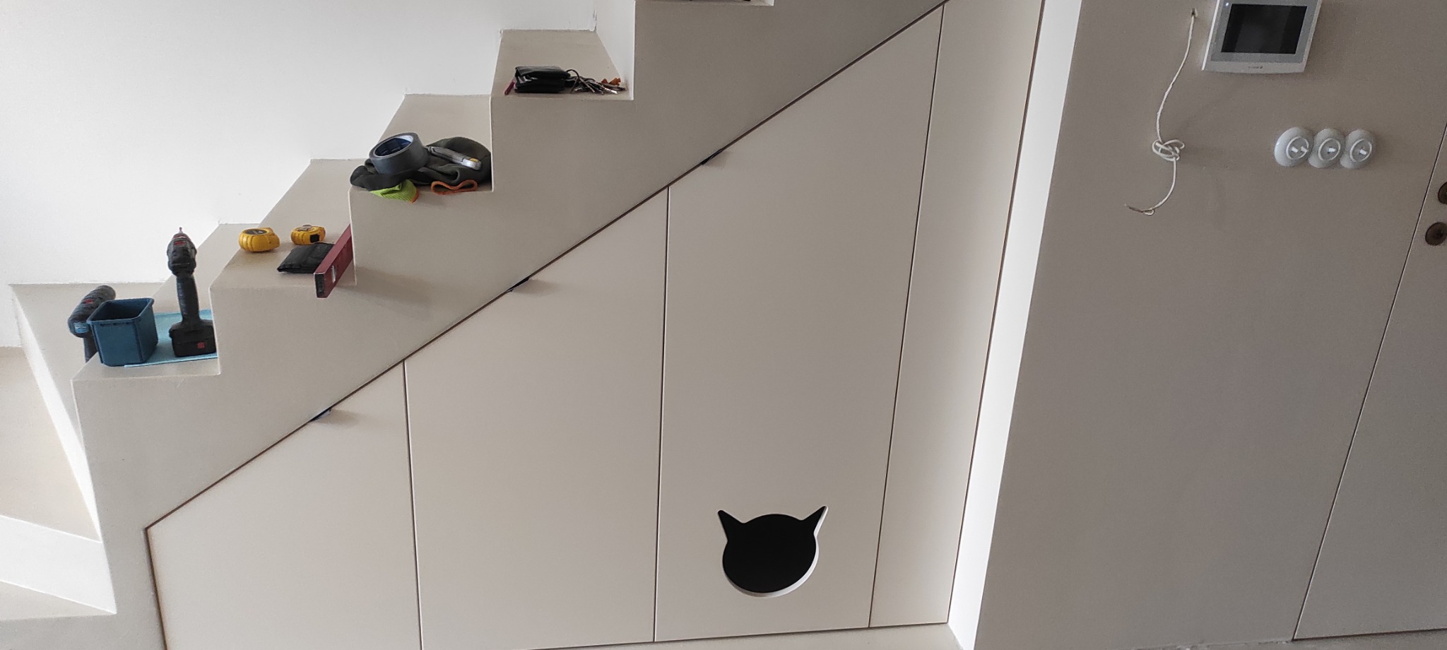 Białe szafki pod schodami z otworem w kształcie kociej głowy. Na schodach narzędzia: wiertarka, poziomica, miarka. Ściana z domofonem i włącznikami.