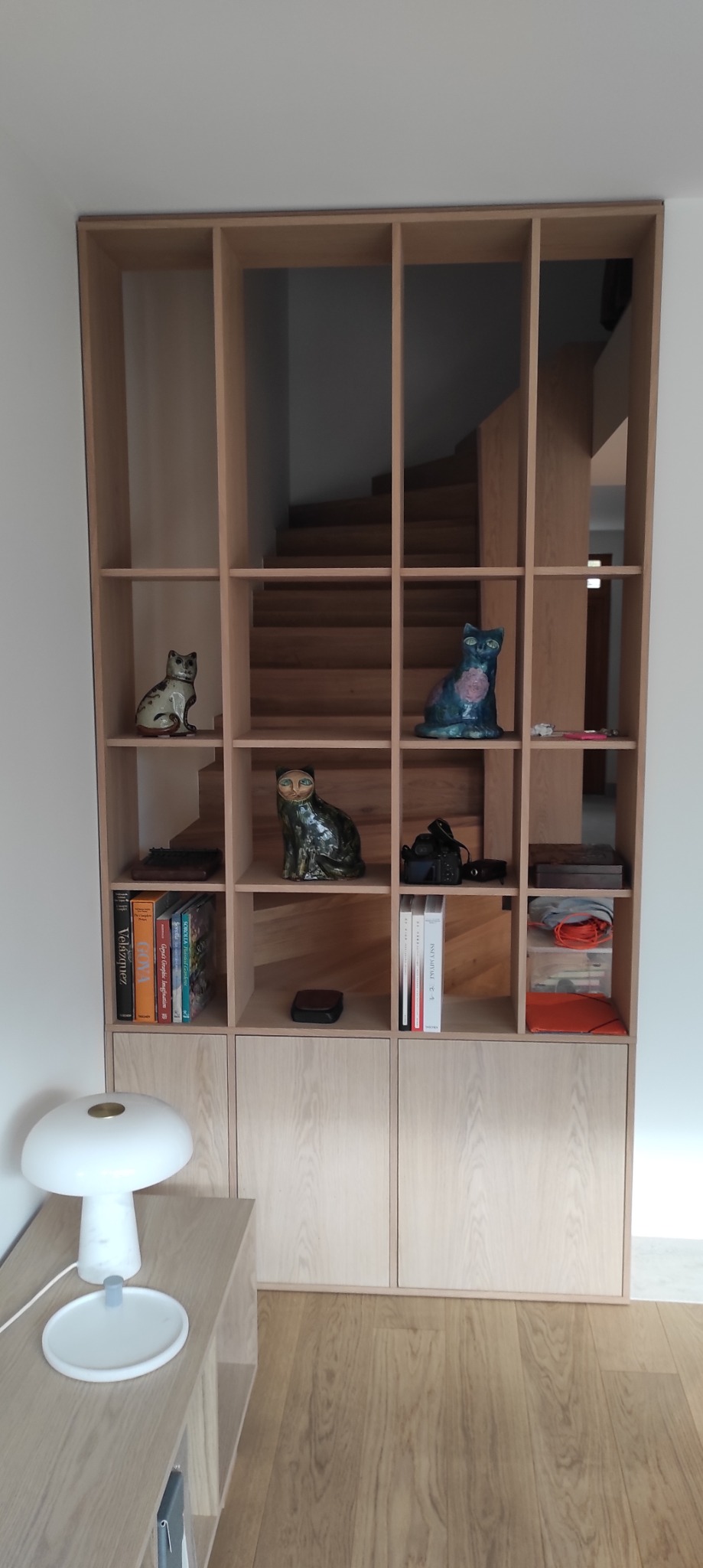 Drewniany regał z półkami i szafkami w jasnym odcieniu drewna, z dekoracjami w postaci figurek kotów. W tle widoczne schody, tworzące spójną całość z meblem.