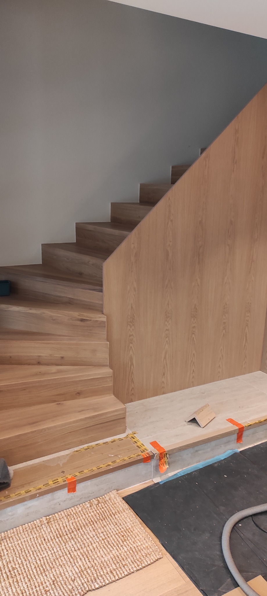 Drewniane schody z panelami ściennymi w trakcie montażu, zabezpieczone taśmą. Widoczne narzędzia i dywanik. Ujęcie z dołu, podkreślające detale wykończenia.