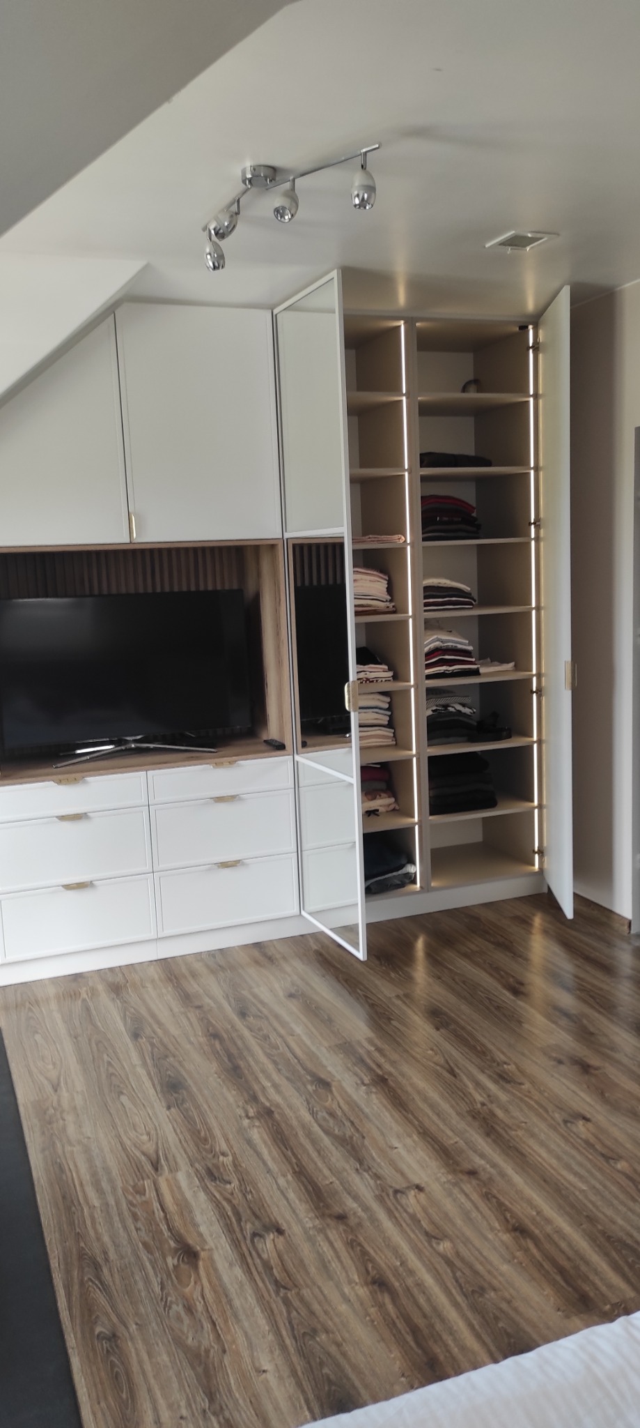 Biała szafa wnękowa z oświetleniem LED i lustrzanymi drzwiami, wkomponowana w skos dachu, z drewnianą podłogą i telewizorem w zabudowie.