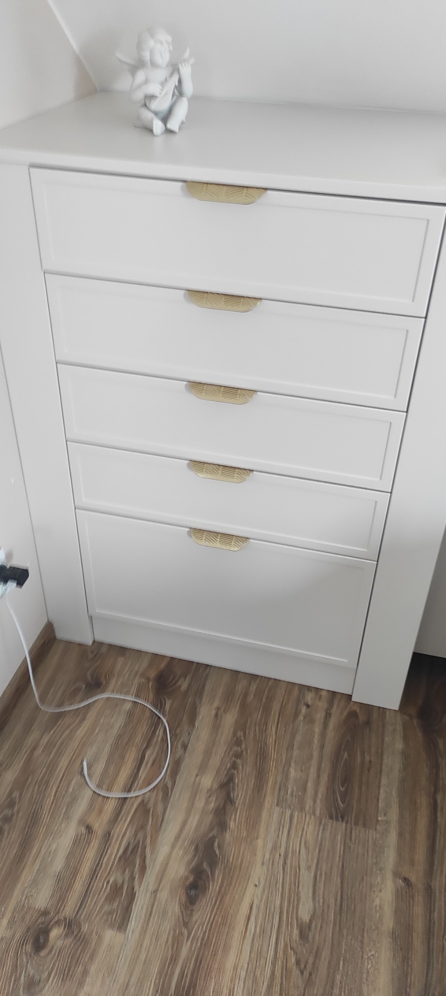 Biała komoda z pięcioma szufladami i dekoracyjnymi uchwytami w kształcie liści. Na komodzie stoi figurka anioła. Widoczna podłoga z paneli.