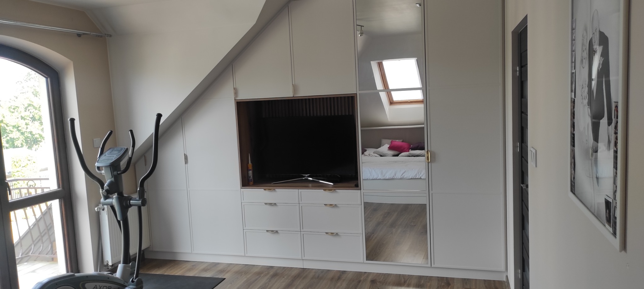 Biała zabudowa meblowa pod skosem z miejscem na TV i lustrzanymi drzwiami, odbijającymi sypialnię. Na pierwszym planie orbitrek.