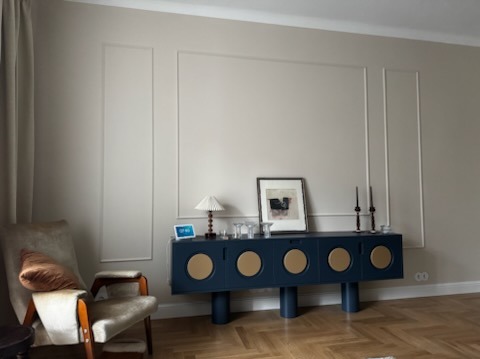 Stylowy salon z granatową komodą z okrągłymi, złotymi akcentami, lampą i dekoracjami. Na podłodze parkiet w jodełkę. Ściany zdobione sztukaterią.
