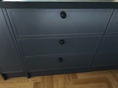 Szczegółowe ujęcie szuflad kuchennych w odcieniach szarości z ciemnymi, okrągłymi uchwytami na tle parkietu ułożonego w jodełkę. Minimalistyczny design.