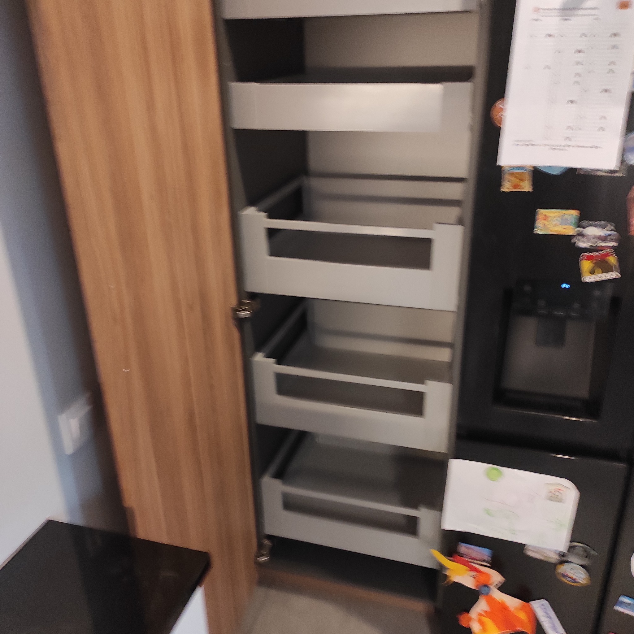 Wysoka szafka kuchenna z drewnianym frontem i wysuwanymi, metalowymi koszami. Widoczne magnesy i rysunki na lodówce obok. Ujęcie z bliska, wnętrze szafki dobrze oświetlone.