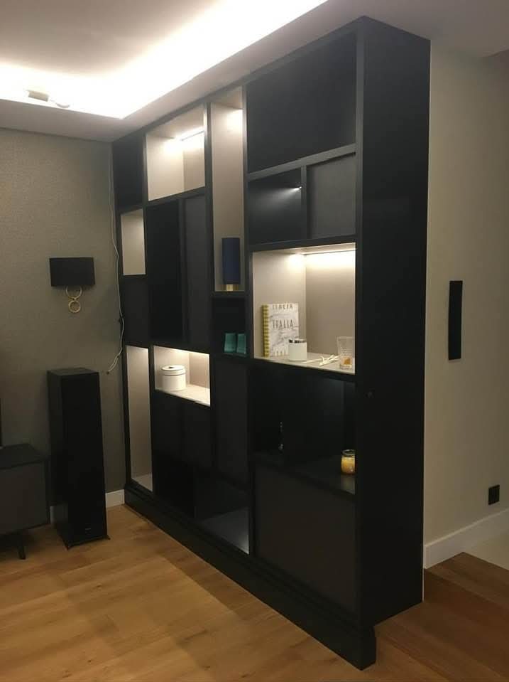 Czarna, nowoczesna szafa wnękowa z podświetlanymi półkami, widoczna z perspektywy podłogi, na której leżą panele. W tle szara ściana z lampą.