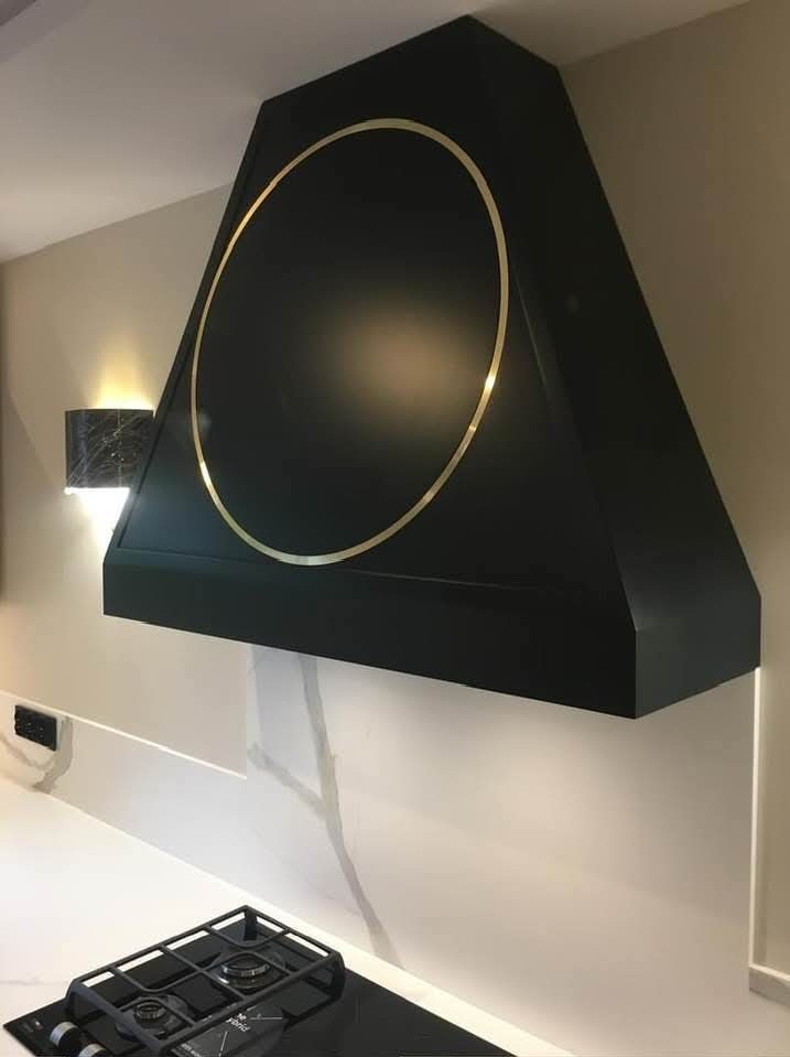 Czarny, trapezowy okap kuchenny z złotym okręgiem, zamontowany nad płytą grzewczą. Minimalistyczny design, oświetlenie punktowe, biały blat z szarymi żyłami.