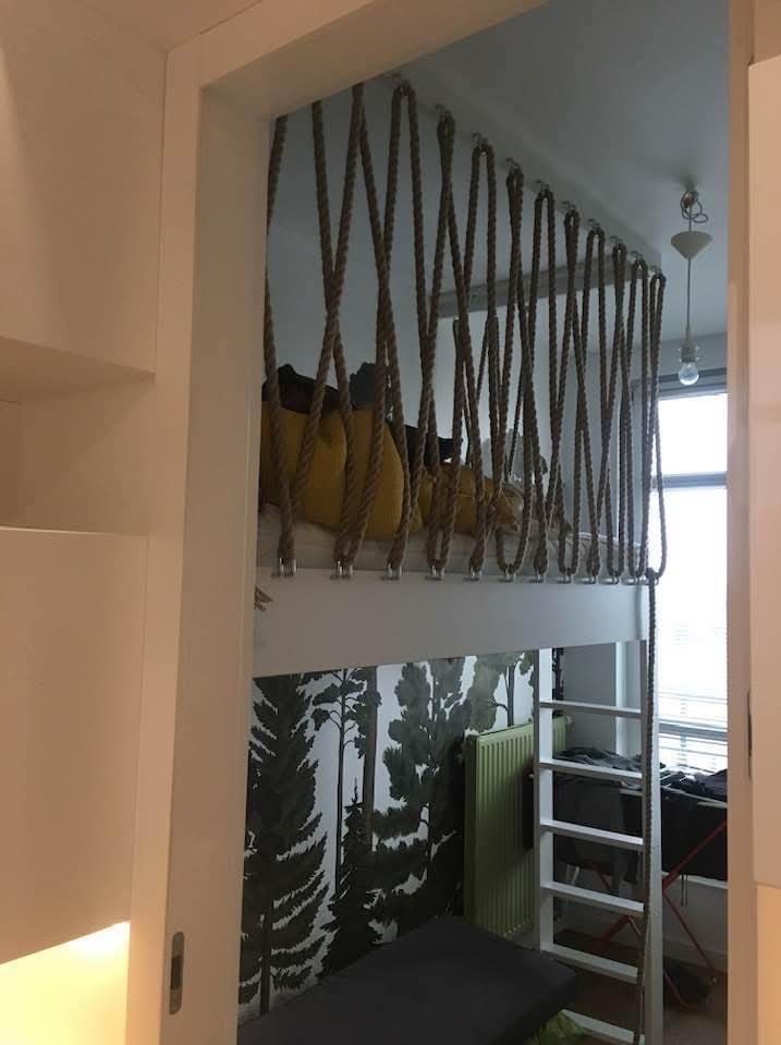 Białe łóżko na antresoli z linową balustradą i drabinką, tapeta z motywem leśnym w pokoju dziecięcym. Nietypowe zabezpieczenie łóżka sznurami.