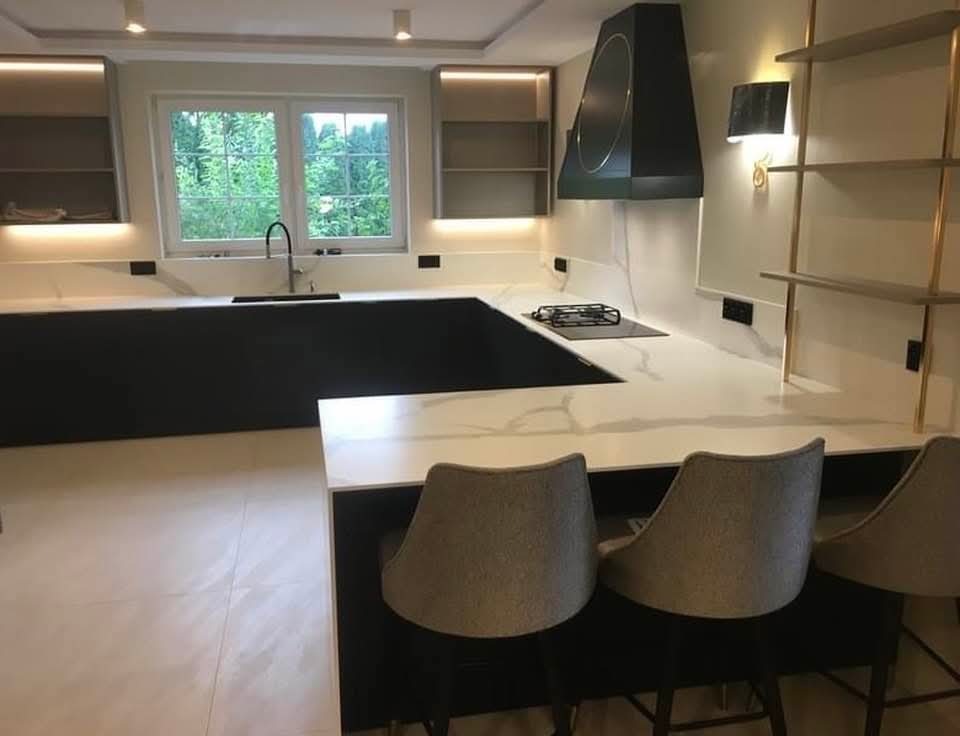 Nowoczesna kuchnia z czarnymi i białymi elementami, oświetleniem LED i marmurowym blatem. Widoczne krzesła barowe i okno z widokiem na zieleń.