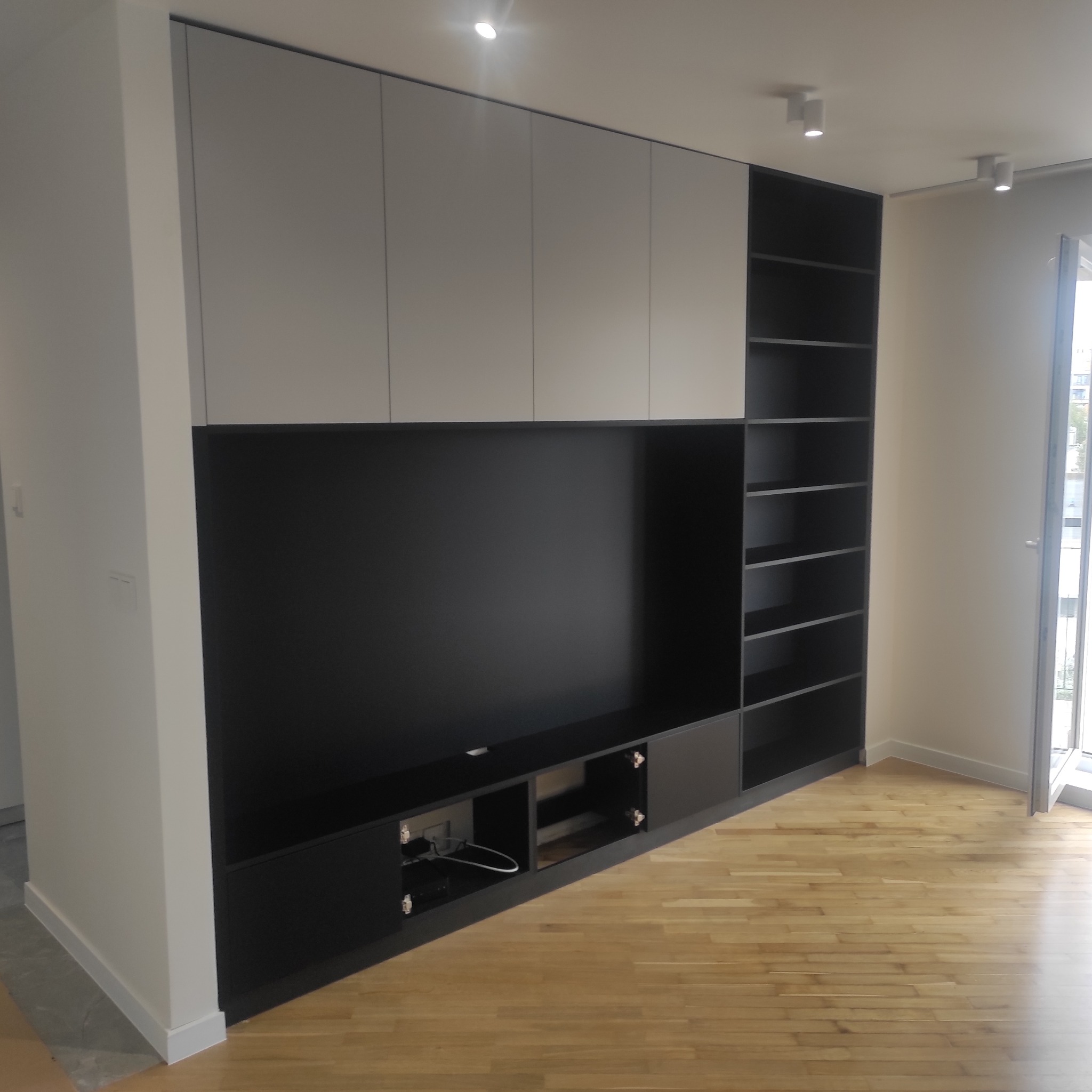 Nowoczesna zabudowa ściany z szafkami w odcieniach szarości i czerni, z otwartymi półkami i miejscem na telewizor, na tle parkietu i jasnych ścian.