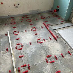 Paweł Petelicki - Układanie płytek podłogowych z użyciem poziomicy i czerwonych klipsów poziomujących w łazience w trakcie remontu. Widoczne instalacje wod-kan.