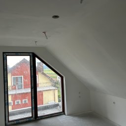 Paweł Petelicki - Poddasze po szpachlowaniu i malowaniu na biało, widoczne okno z brązową ramą i fragment instalacji elektrycznej na suficie. Ściany gładkie, przygotowane do dalszych prac.