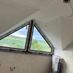 Paweł Petelicki - Poddasze w trakcie wykończenia: trójkątne okno z widokiem na pola, surowe ściany z instalacjami, przygotowane deski podłogowe. Widoczny etap prac wykończeniowych.