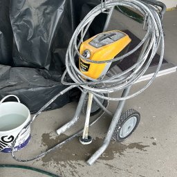 Paweł Petelicki - Profesjonalny agregat malarski Wagner na wózku, z widocznymi śladami farby, obok wiadro z wodą i folia ochronna. Sprzęt gotowy do malowania.