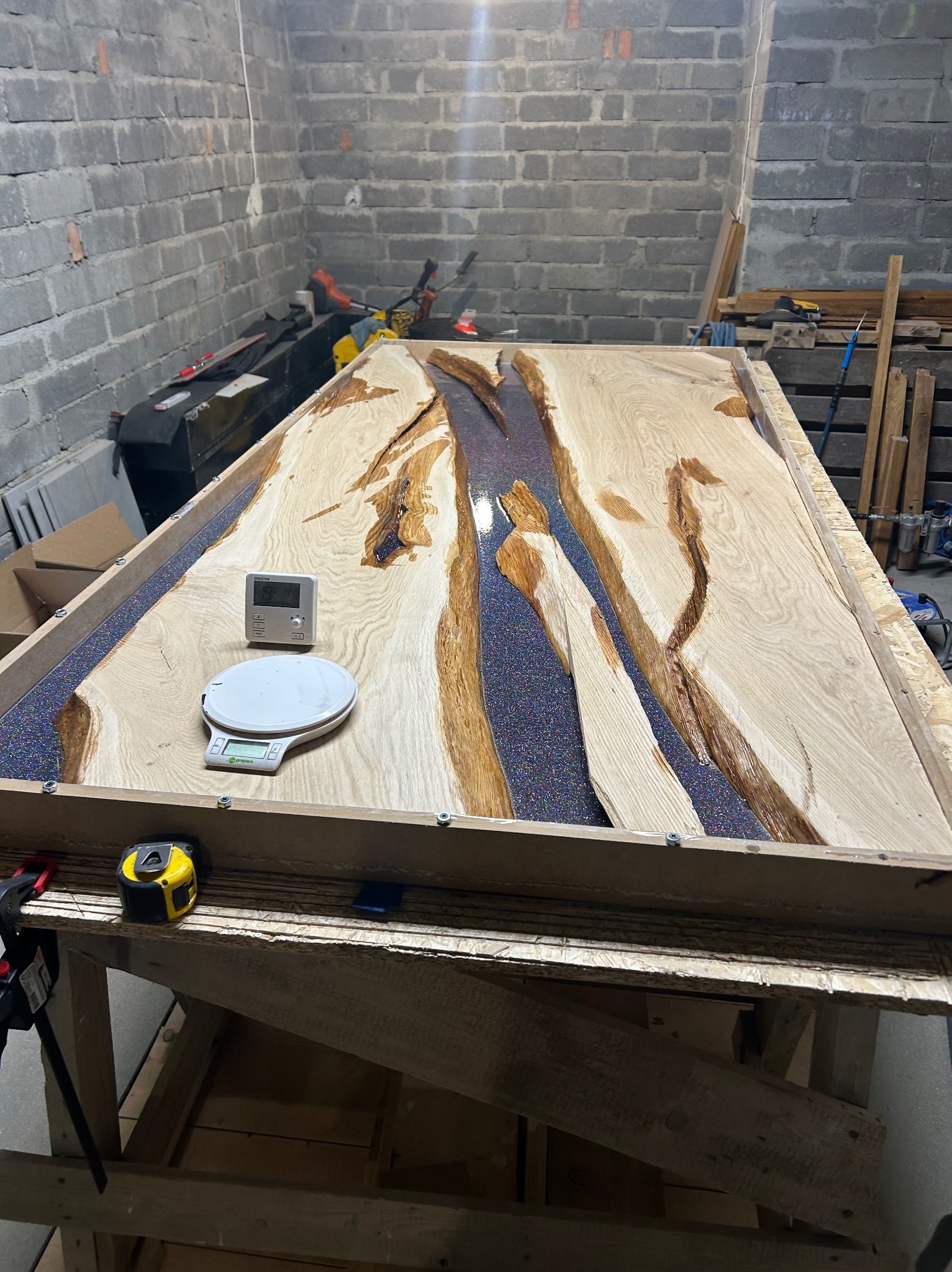 Stół z drewna i żywicy epoksydowej w trakcie tworzenia. Widoczne drewniane elementy, zalewana żywica z brokatem, waga i narzędzia w warsztacie.