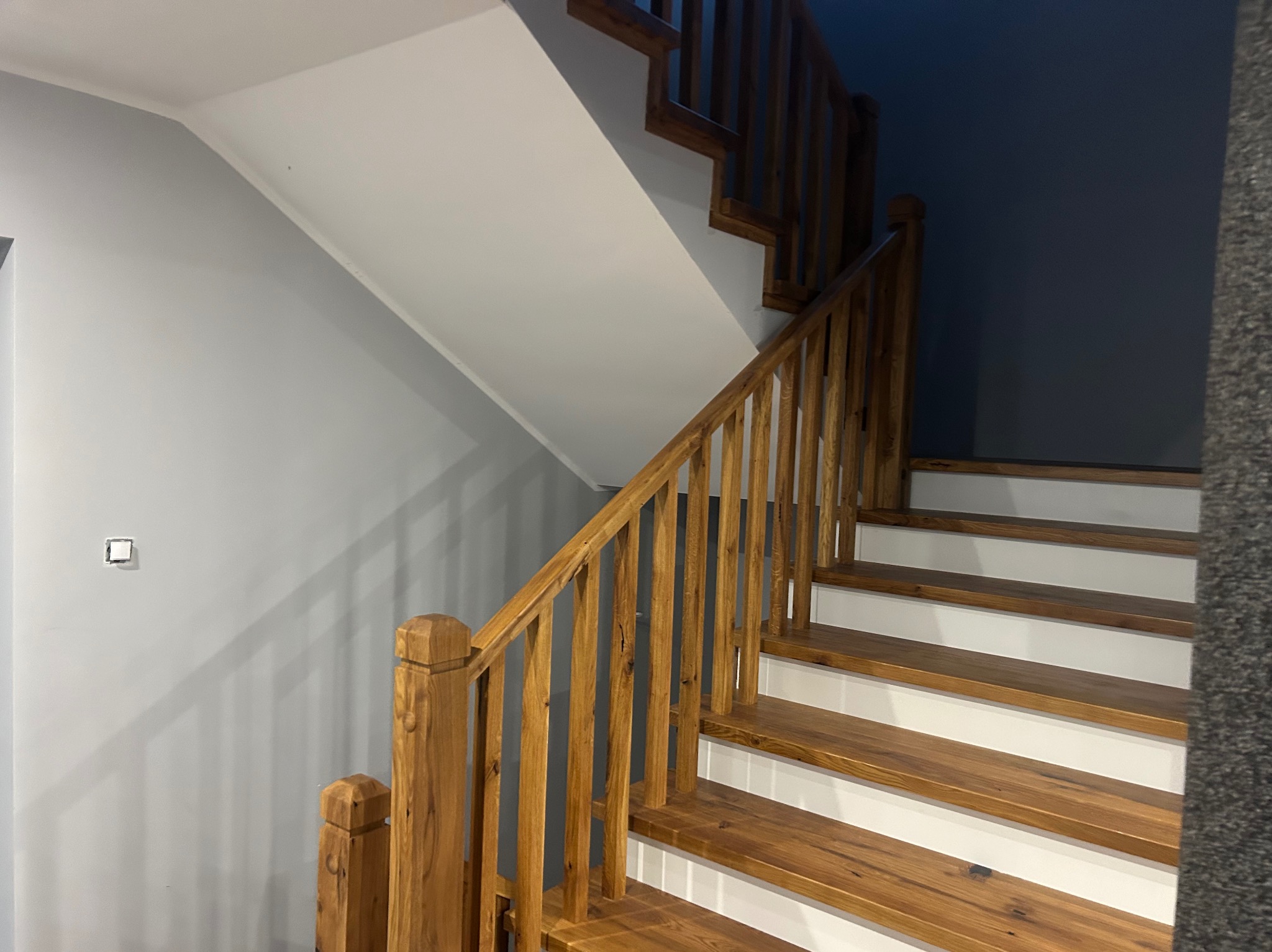 Drewniane schody z białymi podstopnicami w jasnym wnętrzu. Naturalne drewno balustrady i stopni kontrastuje z szarą ścianą i białym sufitem. Widoczny fragment włącznika światła.