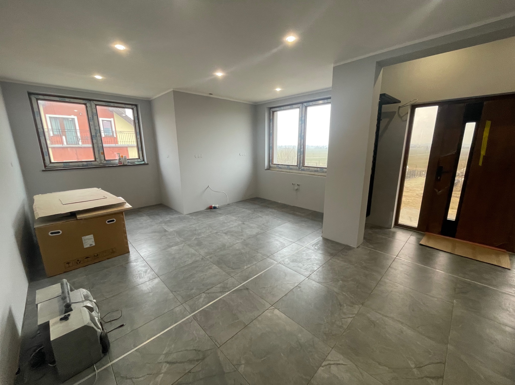 Szare płytki podłogowe w nowym, jasnym pokoju z dwoma oknami i drzwiami wejściowymi. Karton i sprzęt budowlany w rogu. Widok na zewnątrz przez okna.