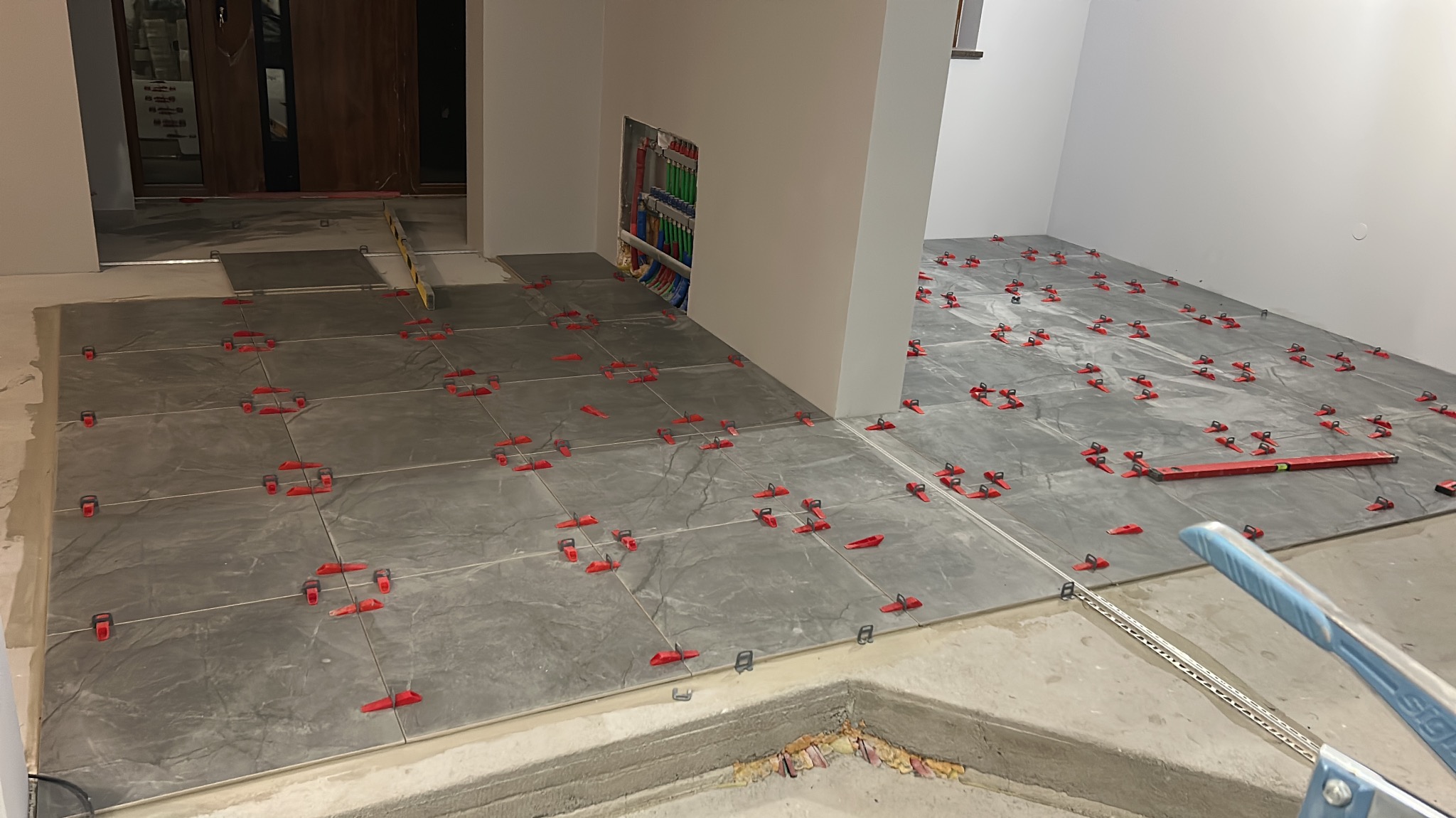 Układanie szarych płytek podłogowych z użyciem czerwonych klipsów poziomujących. Widoczna instalacja elektryczna w ścianie i poziomica. W tle drewniane drzwi.