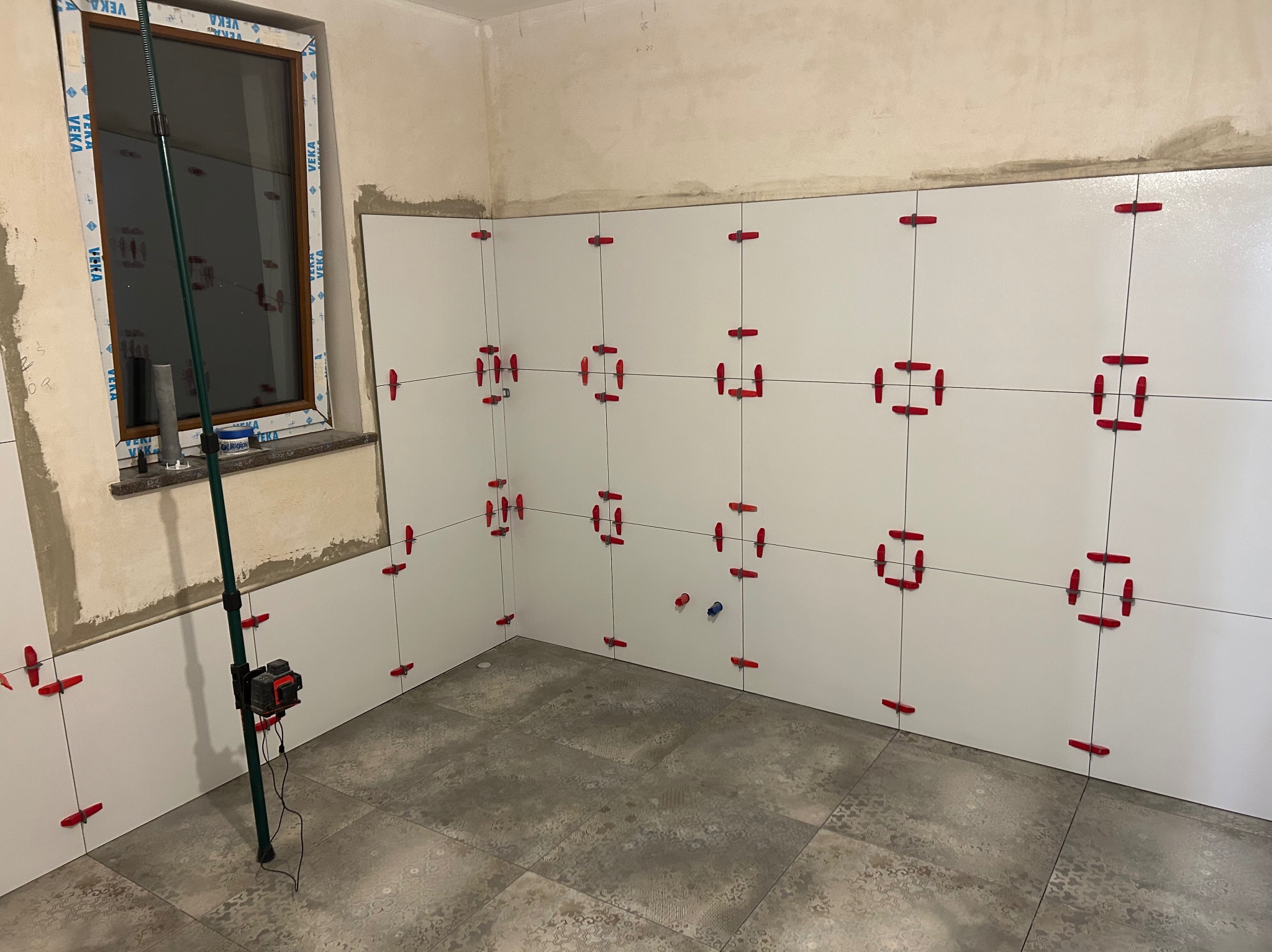 Białe płytki ścienne z czerwonymi krzyżykami dystansowymi w trakcie układania, szare płytki podłogowe, poziomica laserowa przy oknie. Etap prac wykończeniowych w łazience.