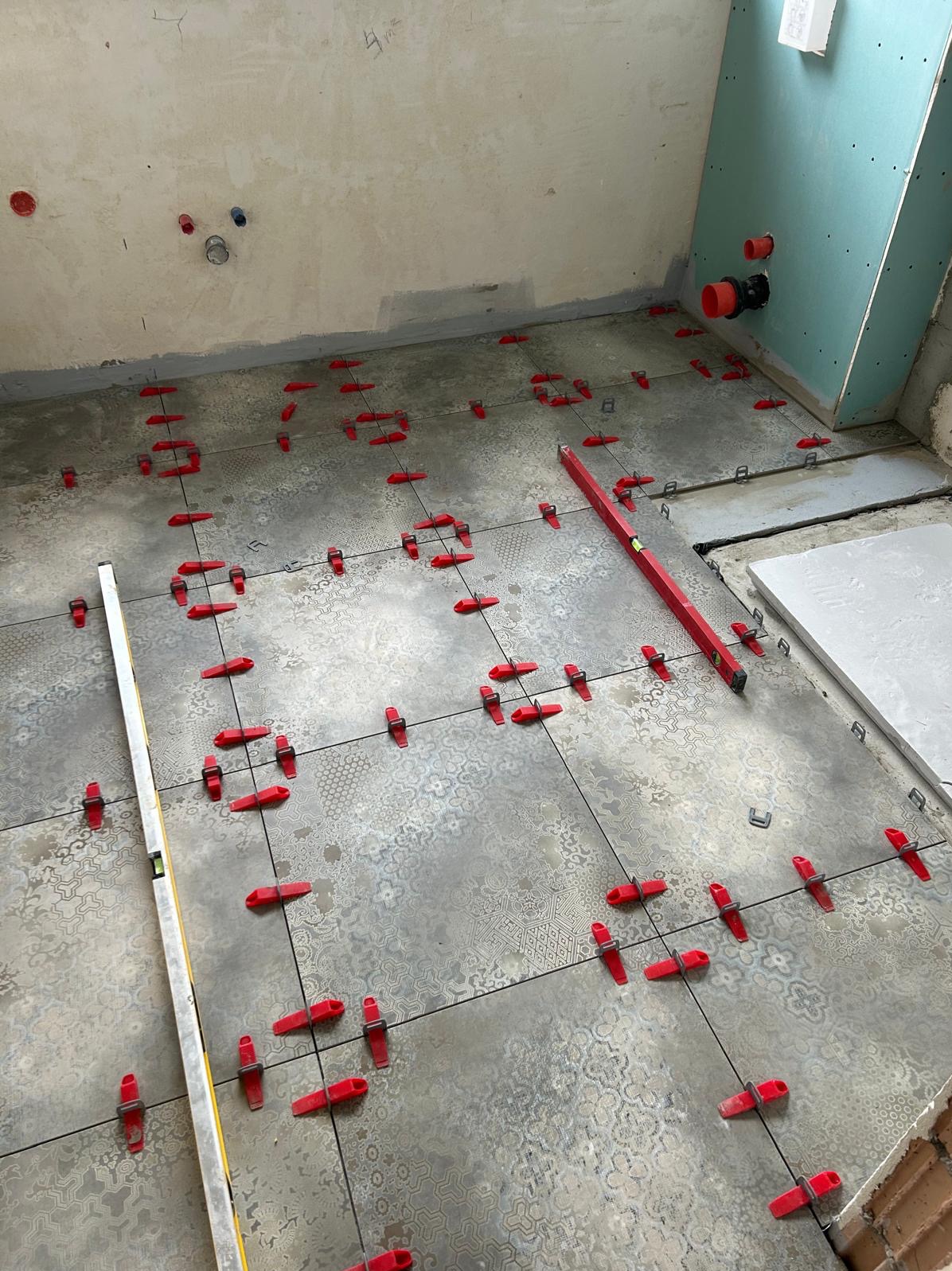 Układanie płytek podłogowych z użyciem poziomicy i czerwonych klipsów poziomujących w łazience w trakcie remontu. Widoczne instalacje wod-kan.