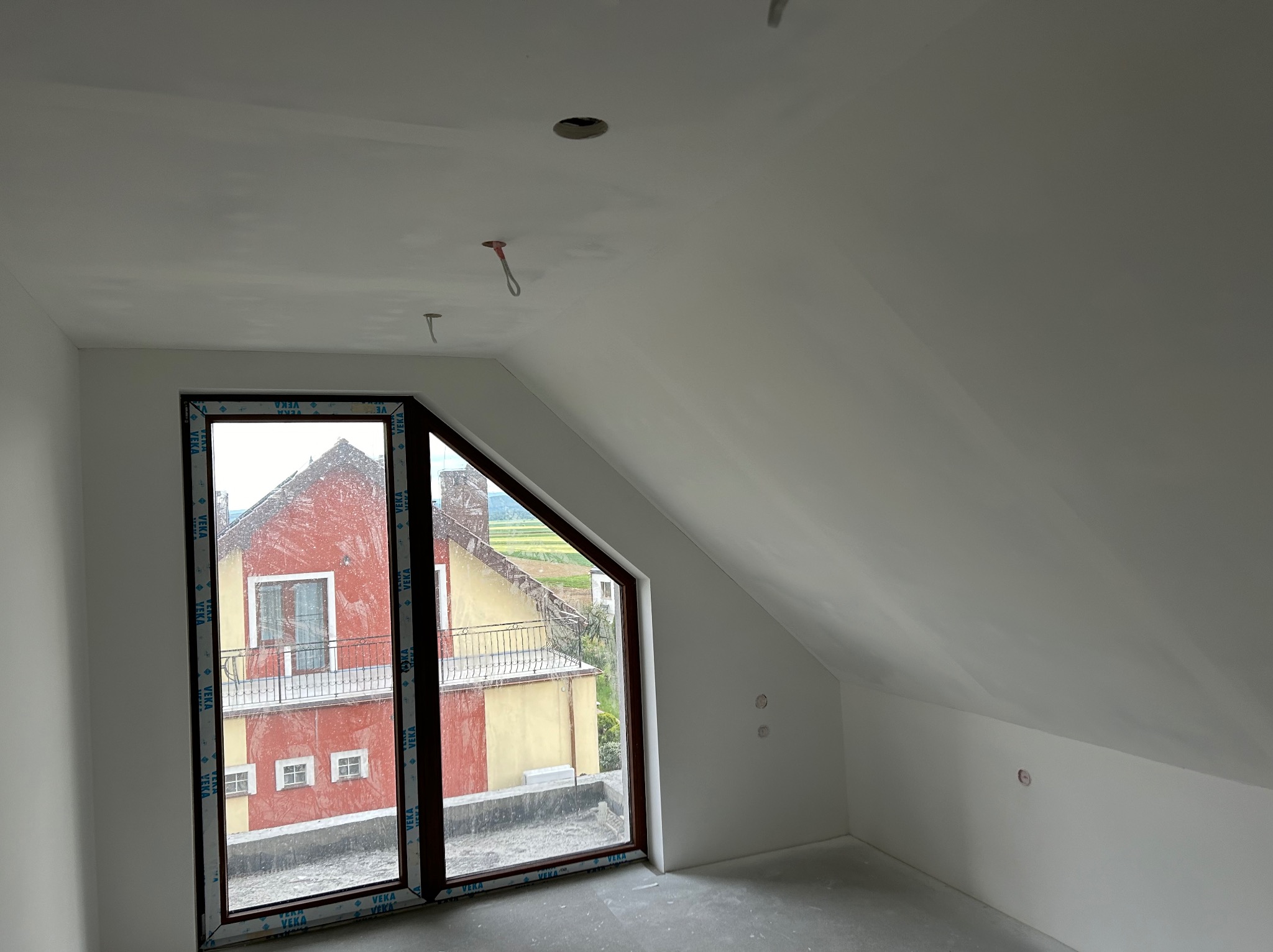 Poddasze po szpachlowaniu i malowaniu na biało, widoczne okno z brązową ramą i fragment instalacji elektrycznej na suficie. Ściany gładkie, przygotowane do dalszych prac.