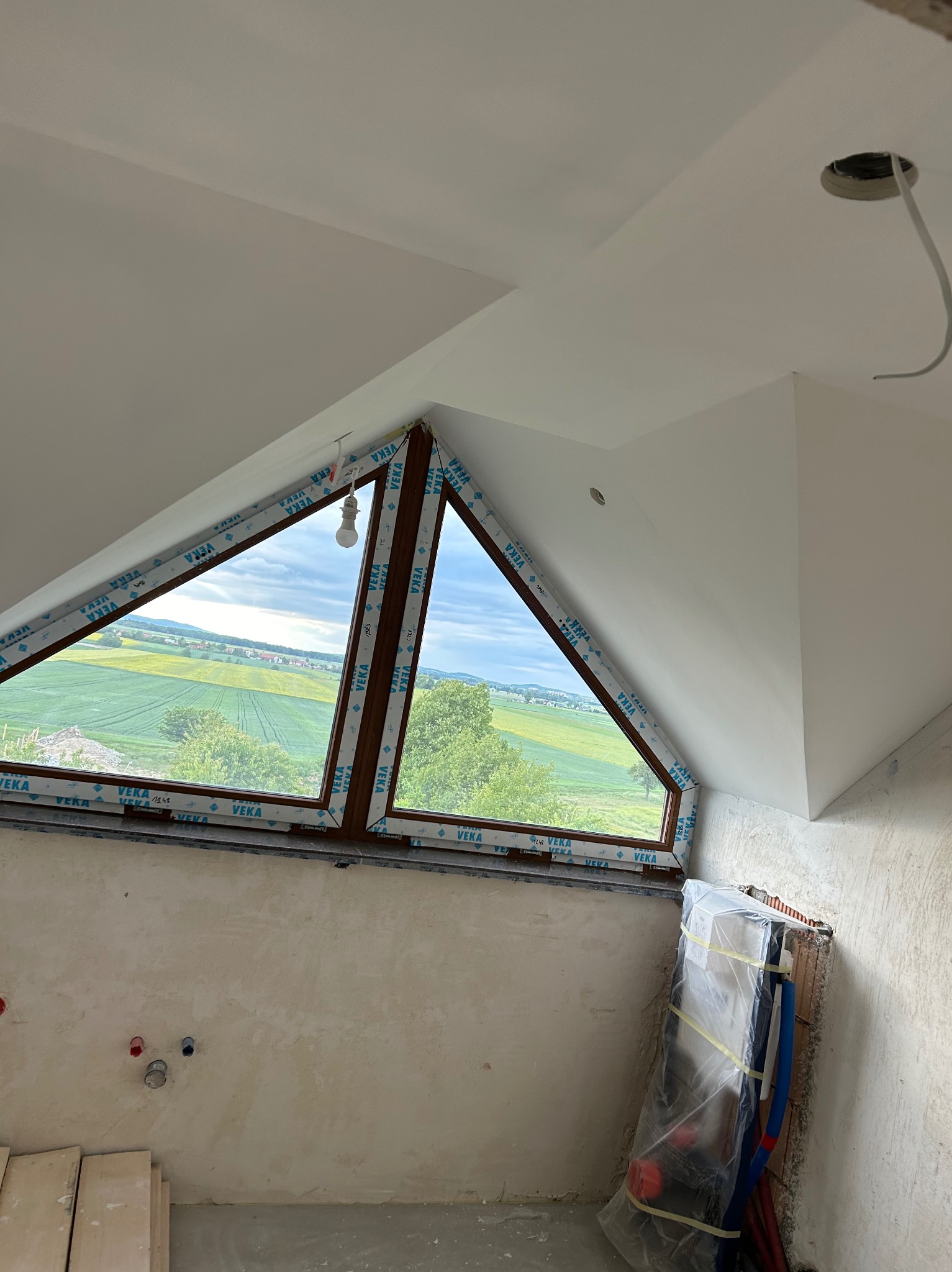 Poddasze w trakcie wykończenia: trójkątne okno z widokiem na pola, surowe ściany z instalacjami, przygotowane deski podłogowe. Widoczny etap prac wykończeniowych.