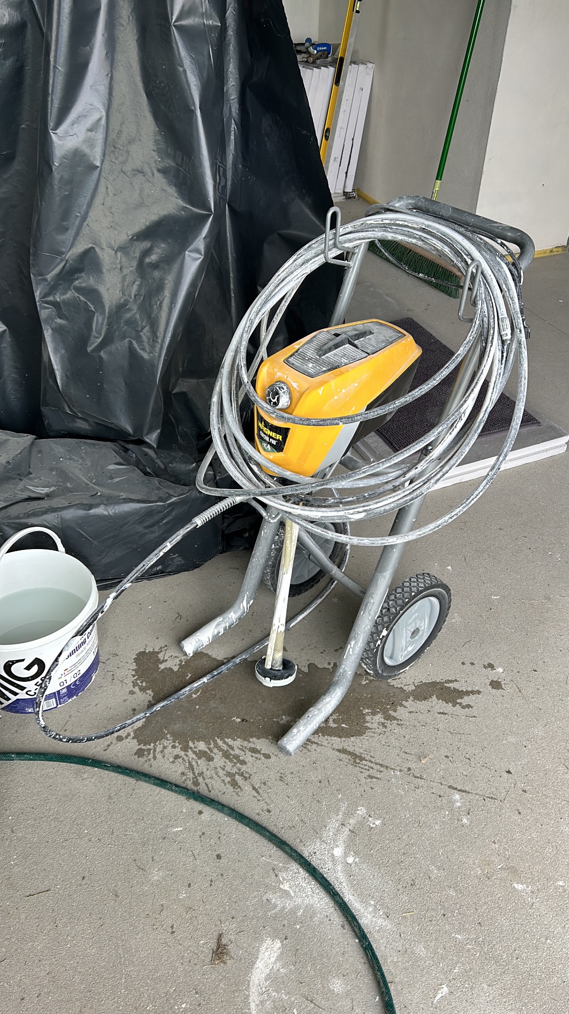 Profesjonalny agregat malarski Wagner na wózku, z widocznymi śladami farby, obok wiadro z wodą i folia ochronna. Sprzęt gotowy do malowania.