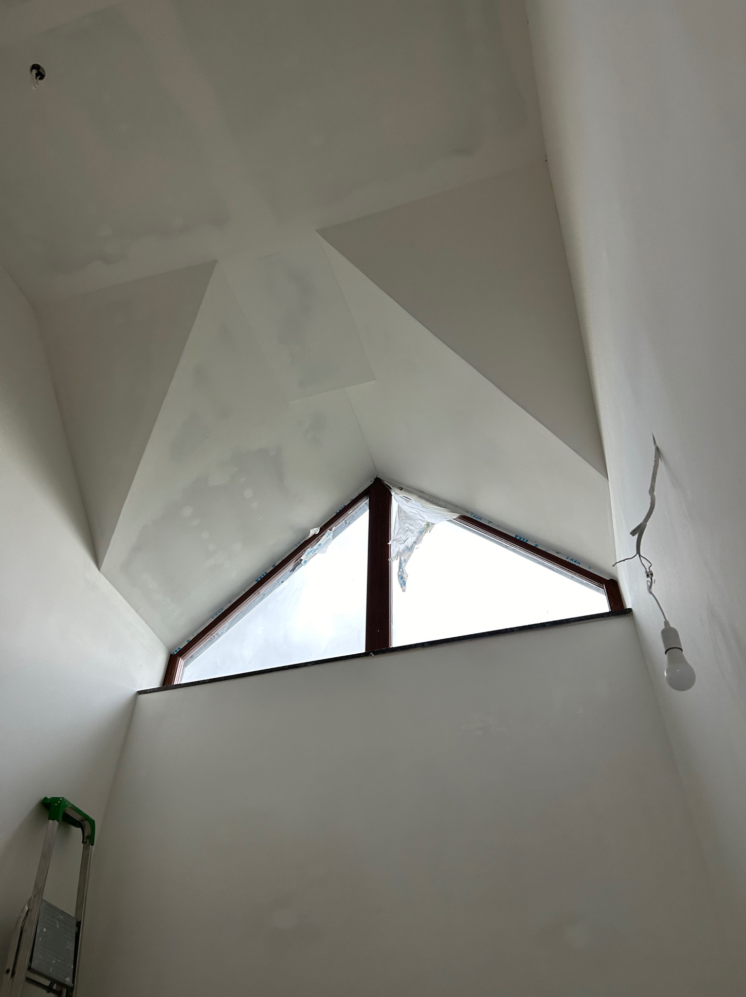 Ściana i sufit po szpachlowaniu, widoczne łączenia płyt gipsowych, okno trójkątne, drabina malarska w rogu. Przygotowanie do malowania.