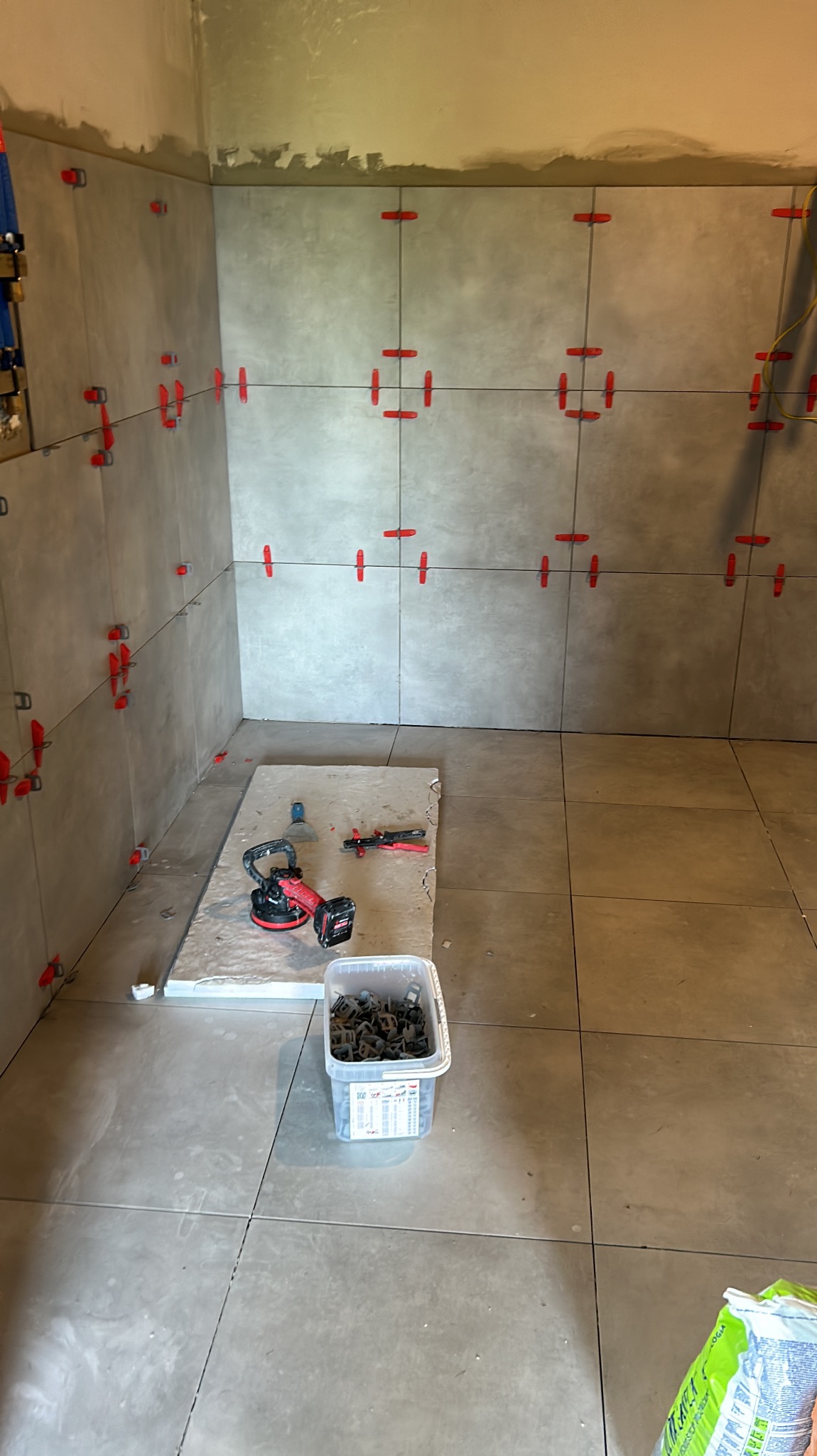 Układanie szarych płytek w łazience w Grochotowie. Widoczne narzędzia glazurnicze i krzyżyki dystansowe. Częściowo wykończona ściana i podłoga.