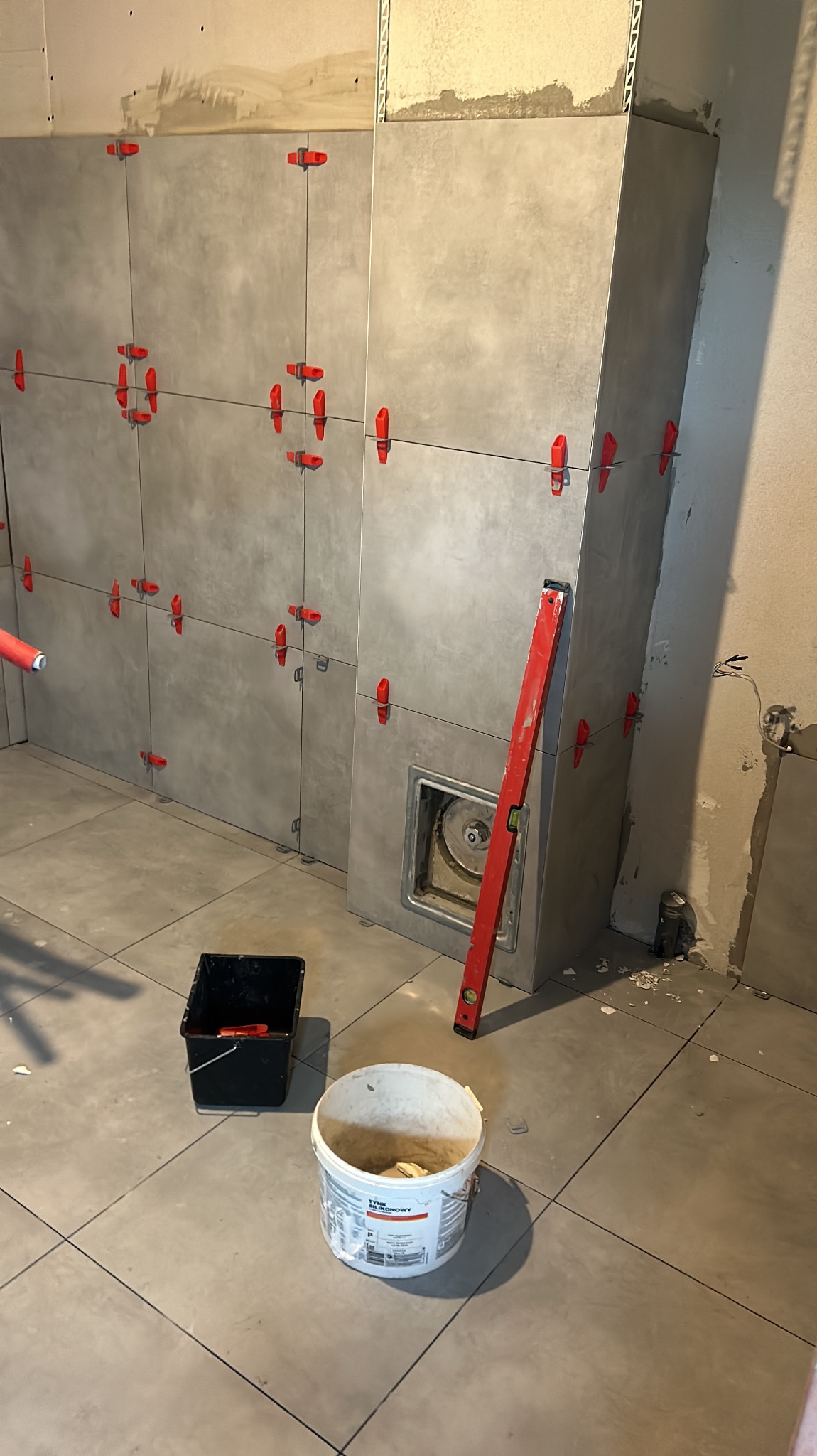 Szare płytki ścienne z czerwonymi krzyżykami dystansowymi, poziomica oparta o ścianę, wiadro z zaprawą i czarne wiaderko na podłodze z płytek. Prace wykończeniowe w łazience.