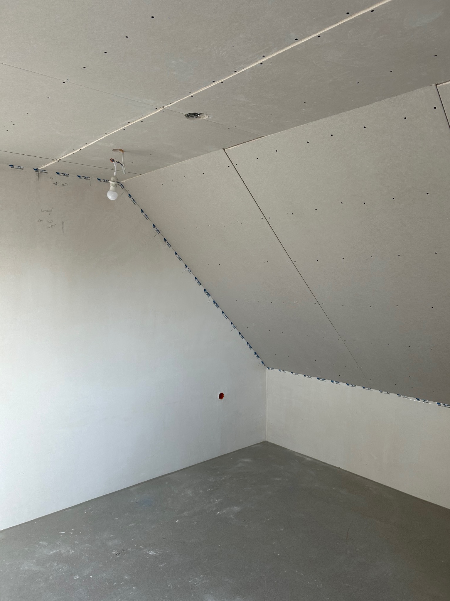 Pokój na poddaszu w trakcie remontu: ściany wyszpachlowane, zamontowane płyty gipsowo-kartonowe na skosach, surowa podłoga. Widoczna żarówka na kablu.