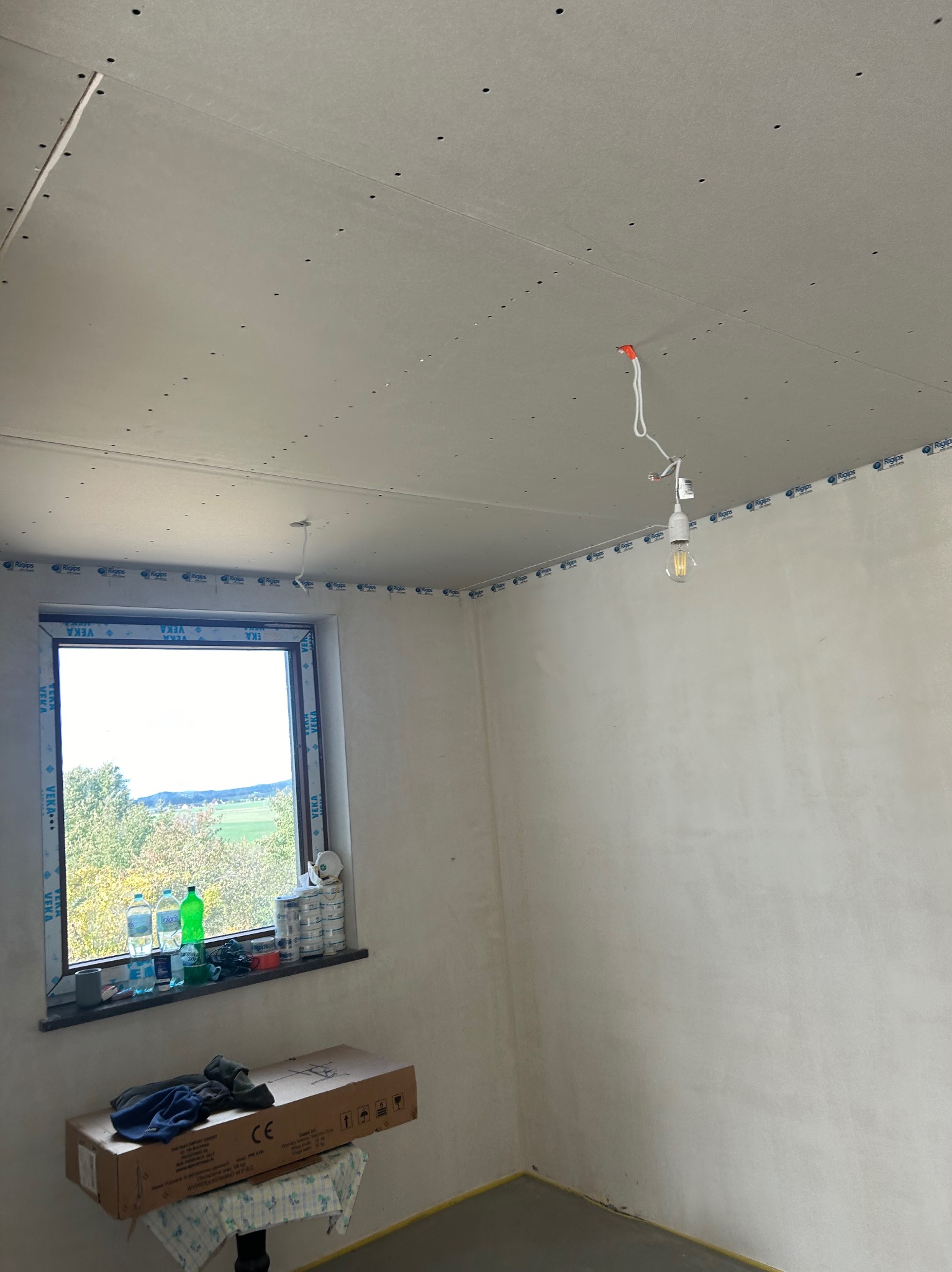 Pokój w trakcie prac wykończeniowych: zamontowane płyty gipsowo-kartonowe na suficie i ścianach, widoczne okno z widokiem na zewnątrz i prowizoryczne oświetlenie.