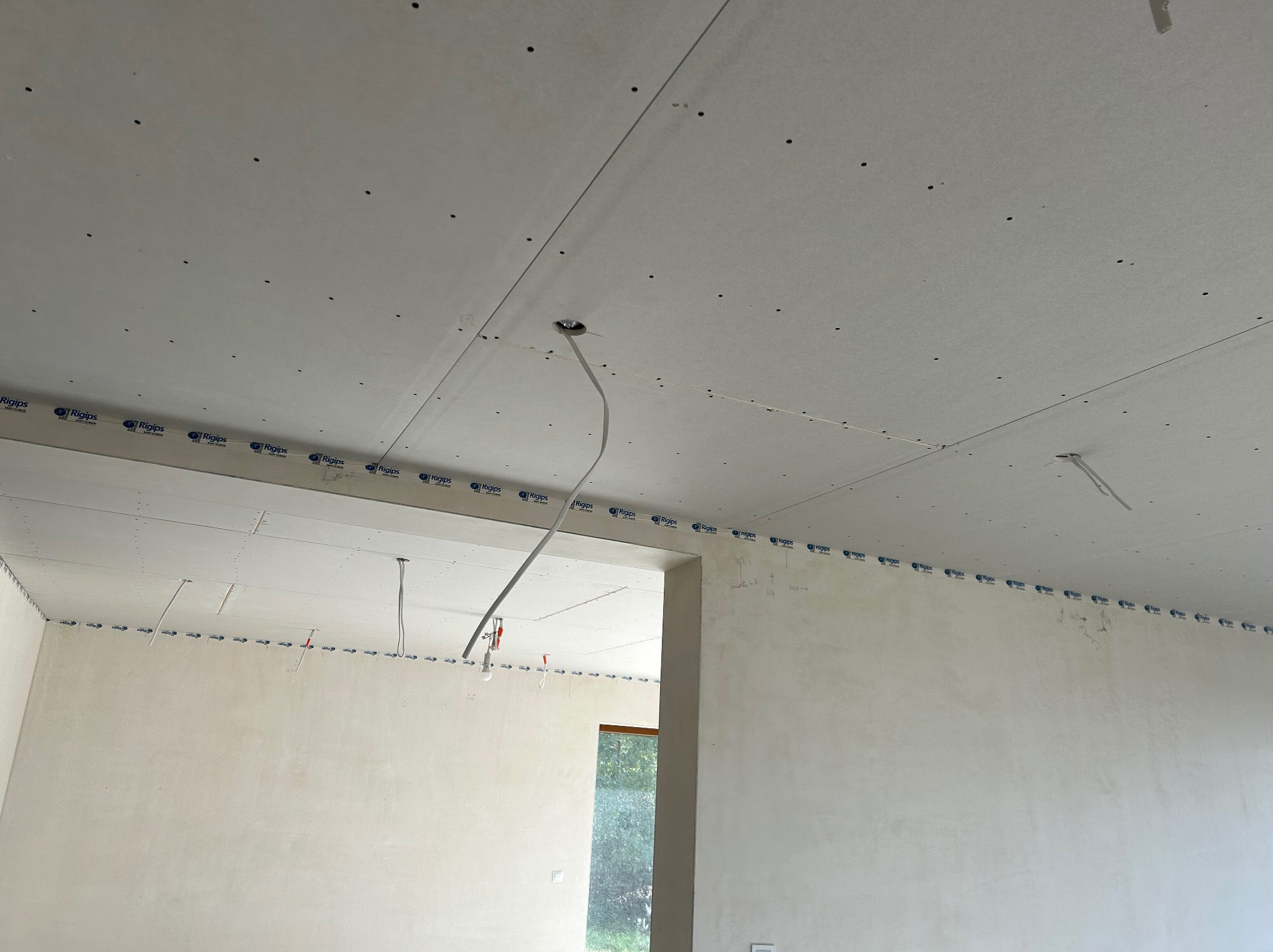 Montaż płyt gipsowo-kartonowych na suficie z widocznymi przewodami elektrycznymi i fragmentem ściany. Widok na surowy stan wykończenia wnętrza.