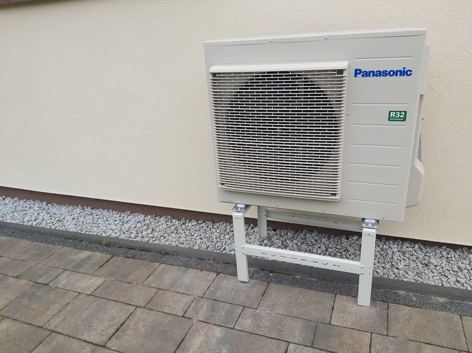 Zamontowana jednostka zewnętrzna pompy ciepła Panasonic R32 na metalowym stelażu przy ścianie budynku, widok z boku na kostce brukowej i kamiennym podsypaniu.