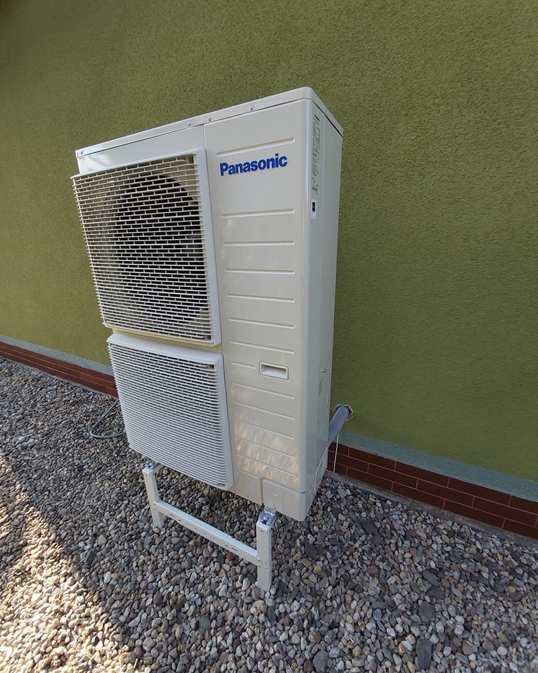 Zewnętrzna jednostka klimatyzacji Panasonic na żwirowym podłożu, wsparta na białym stelażu, przy zielonej ścianie z ceglanym cokołem. Widoczne kratki wentylacyjne.
