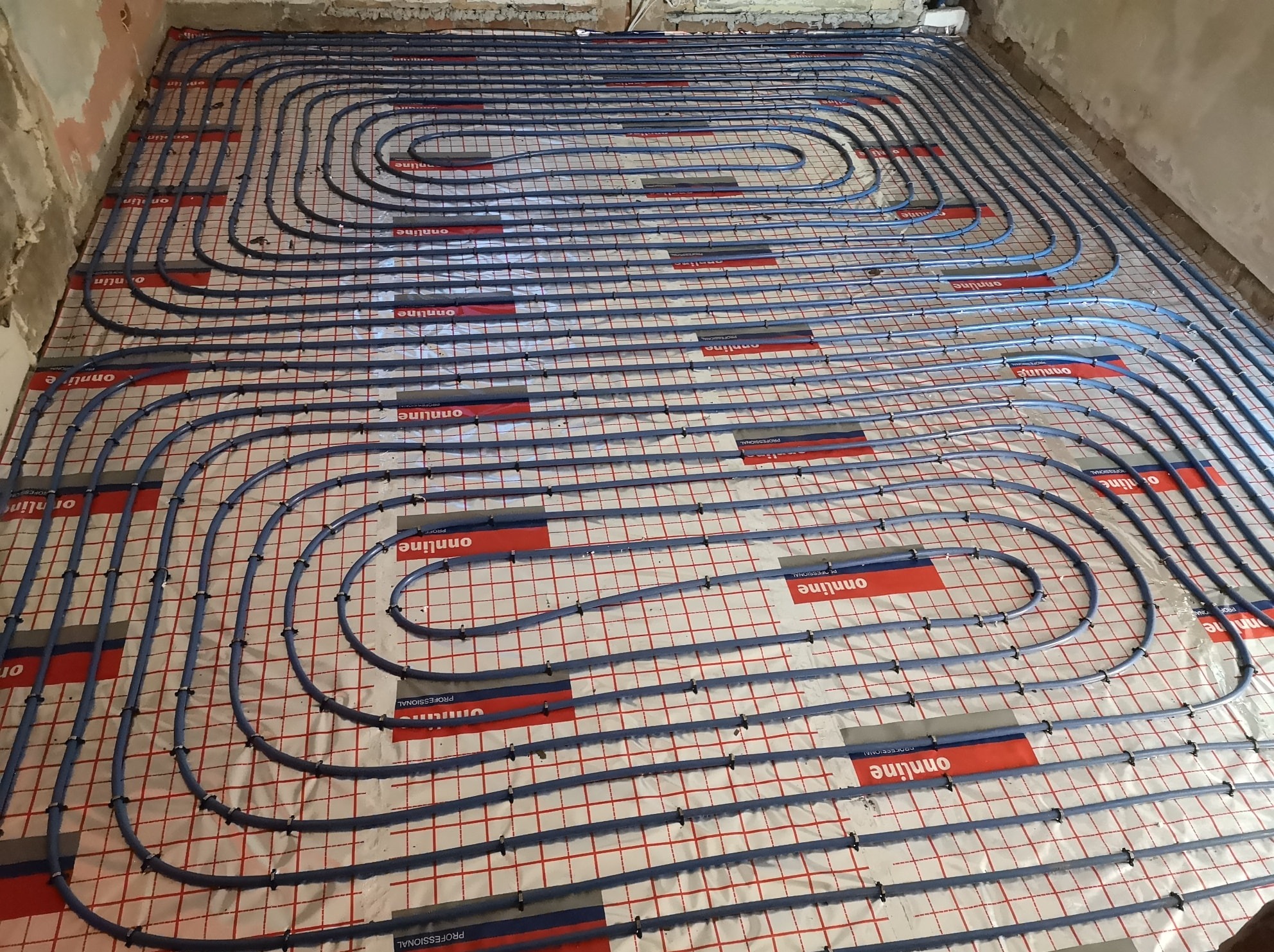 Ułożone rury ogrzewania podłogowego na folii z siatką, gotowe do zalania posadzką. Widoczne mocowania rur. Surowe ściany w tle.
