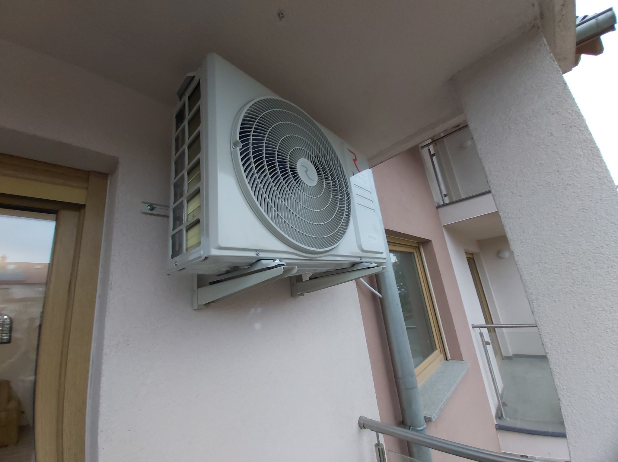 Biały klimatyzator zamontowany na elewacji budynku, widoczny z dołu pod kątem. Na drugim planie okno i fragment balkonu z metalową barierką.