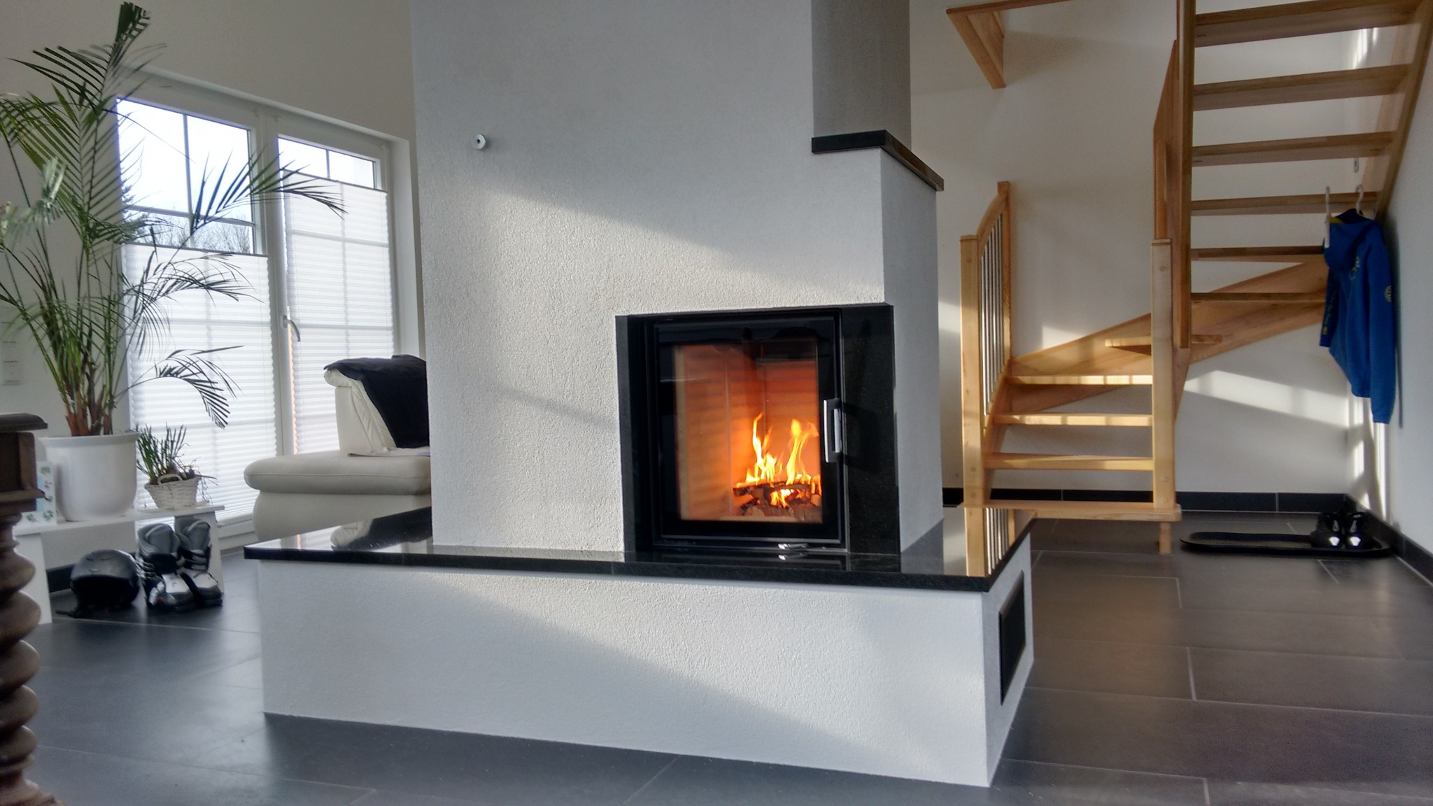 Nowoczesny kominek z czarnym obramowaniem i płomieniami, w jasnym wnętrzu z drewnianymi schodami i roślinami doniczkowymi. Minimalistyczny design.