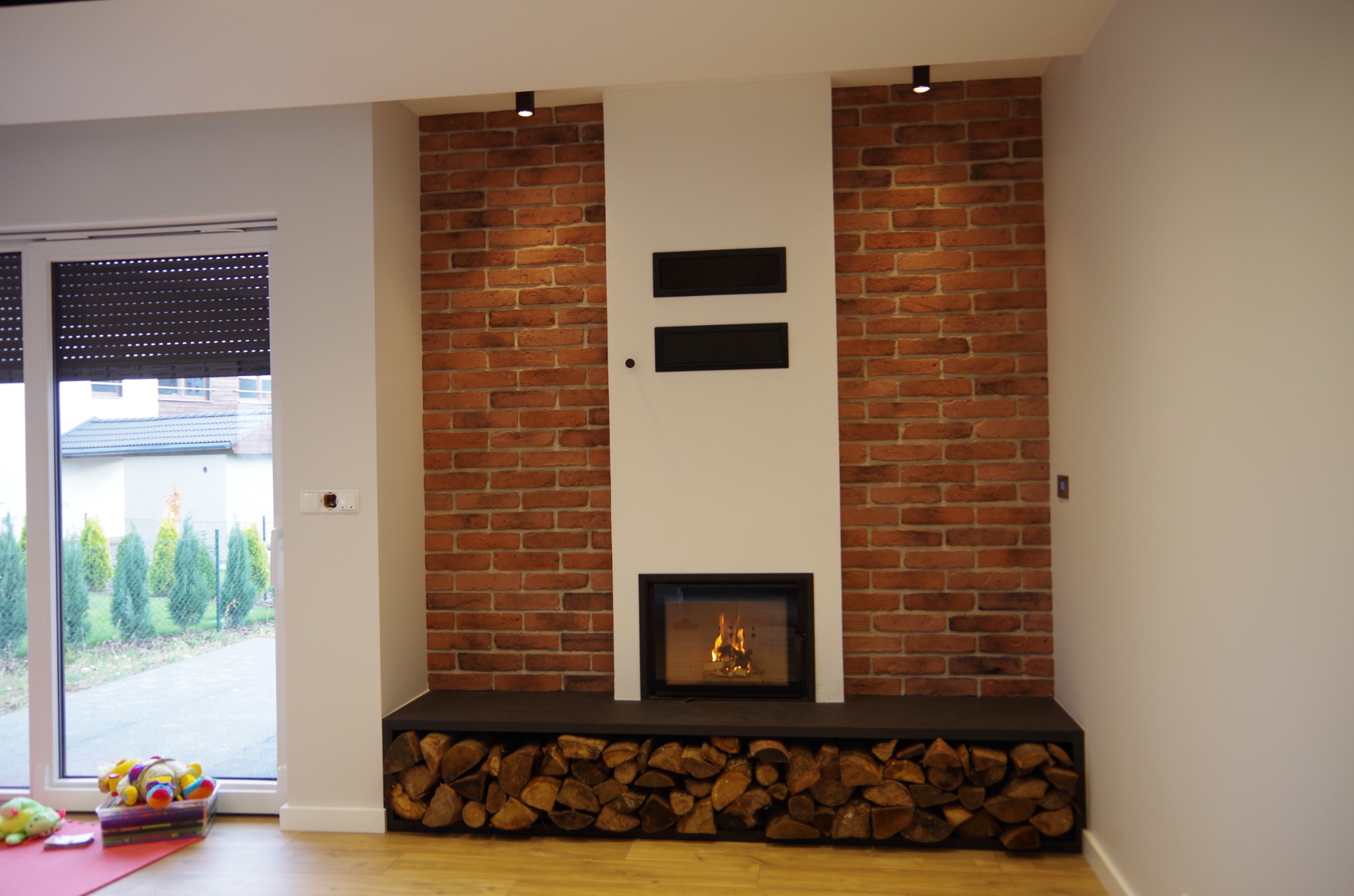 Murowany kominek z cegły z paleniskiem i drewnem u podstawy, oświetlony punktowymi lampami, na tle jasnych ścian i podłogi. Widok z wnętrza domu na ogród.