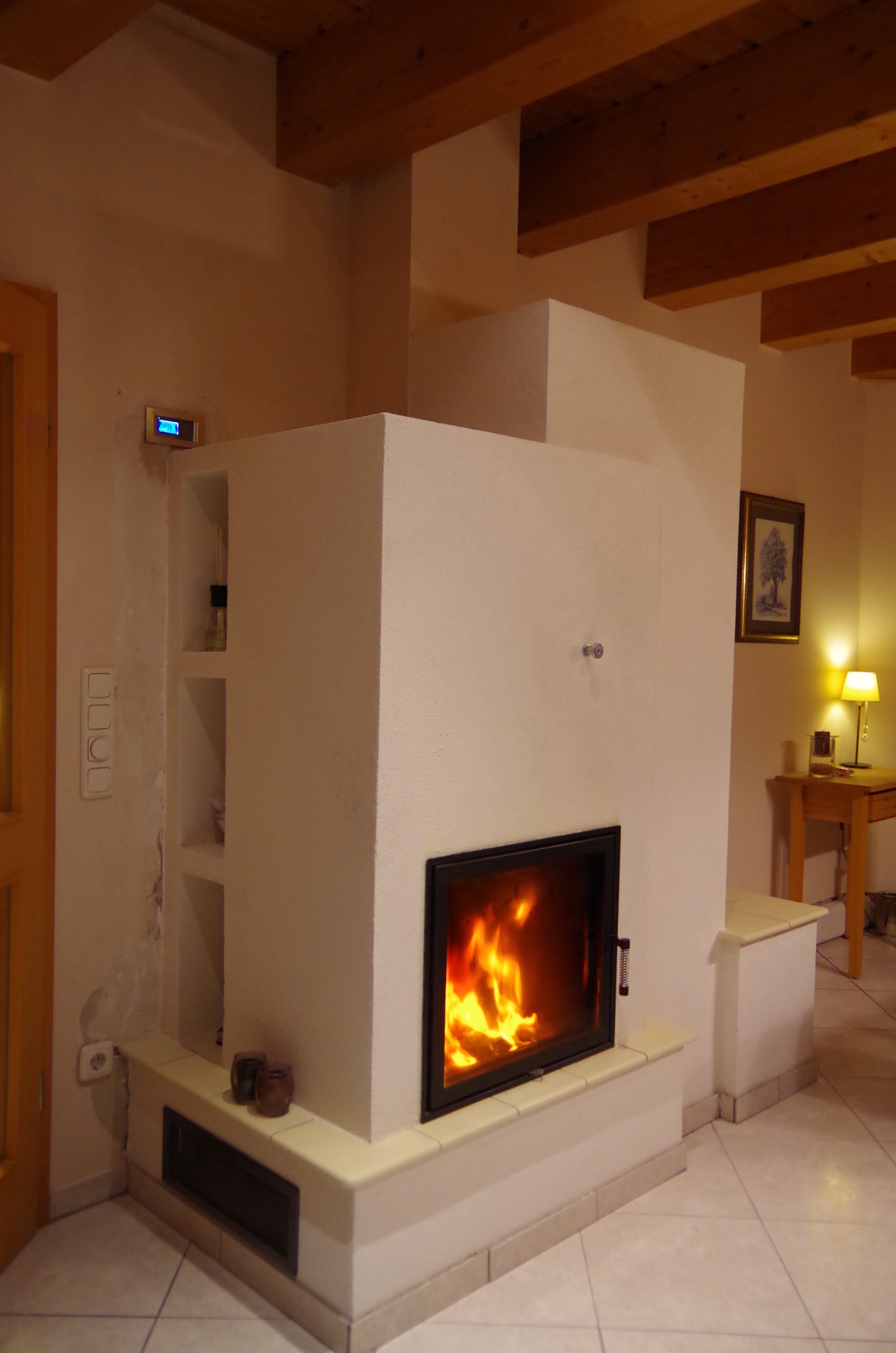 Biały kominek kaflowy z paleniskiem, wbudowany w ścianę, z półkami po lewej stronie. Widoczne płomienie, drewniane belki stropowe i fragment podłogi z płytek.