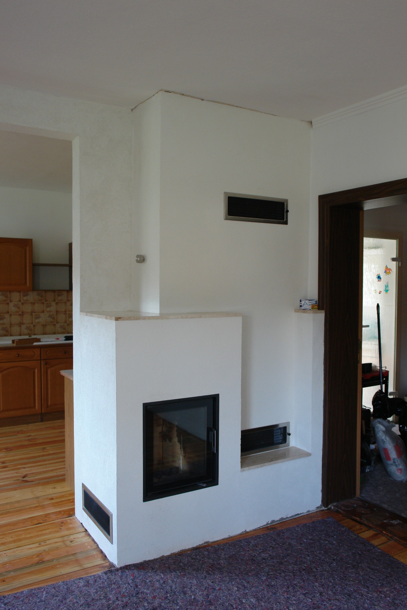 Biały kominek z czarną szybą w salonie, widoczne kratki wentylacyjne, drewniana podłoga i fragment kuchni w tle. Minimalistyczny design kominka.