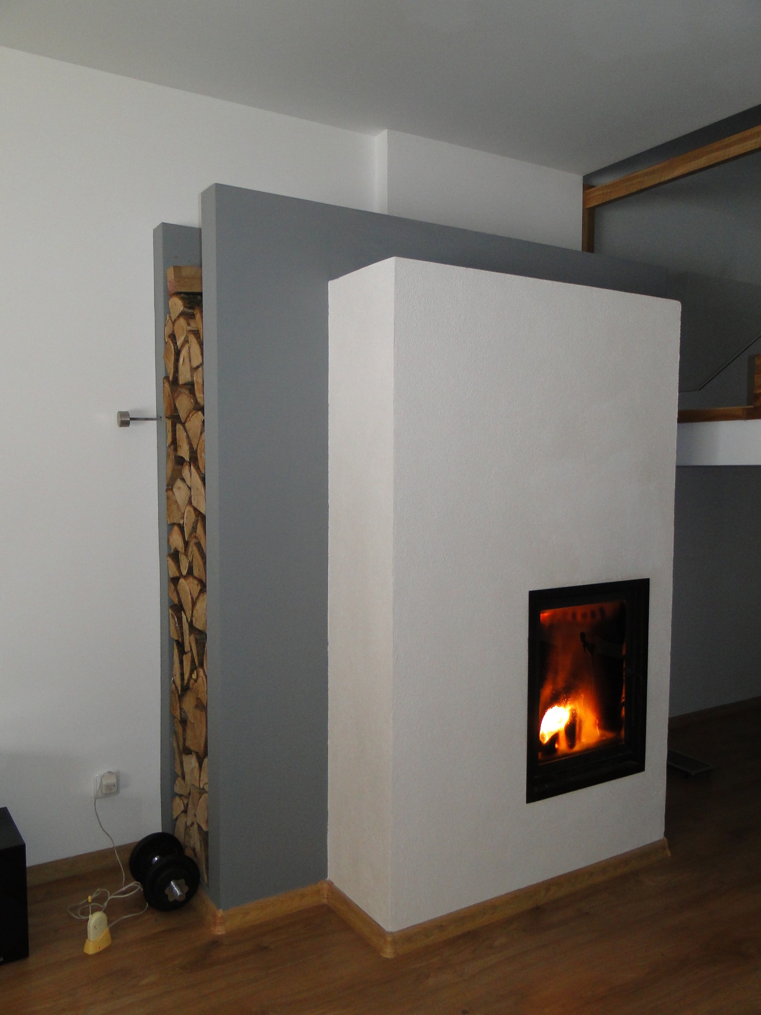 Nowoczesny kominek z obudową w odcieniach bieli i szarości, z wnęką na drewno. Płomienie widoczne przez czarną ramę paleniska, drewniana podłoga.