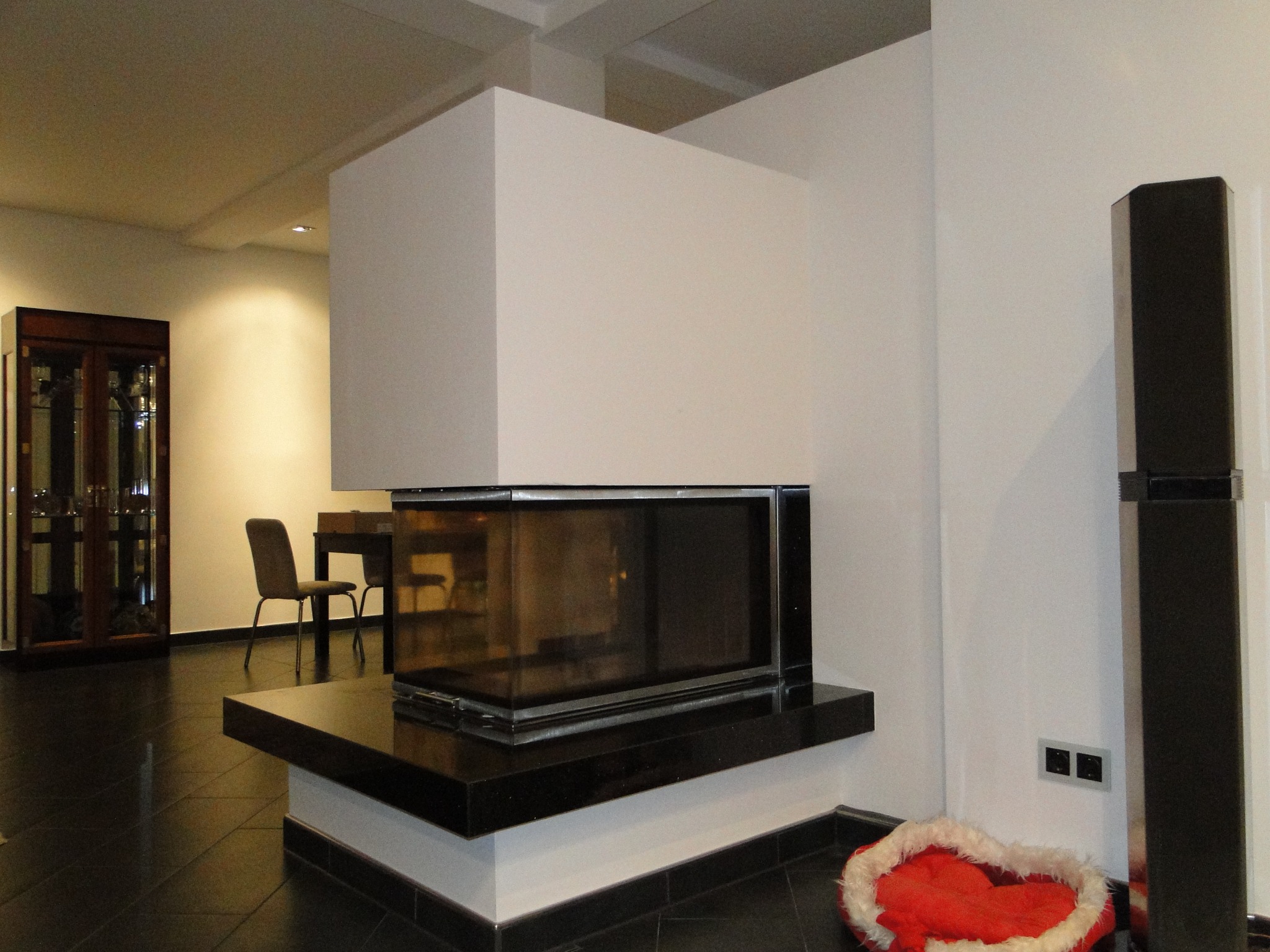 Nowoczesny kominek narożny z czarnym obramowaniem i blatem, w tle widoczne meble i ciemna podłoga. Obok kominka leży czerwone legowisko dla zwierząt.