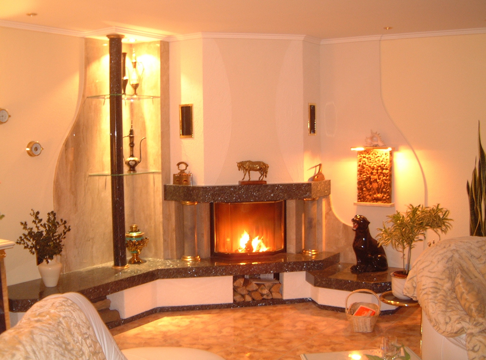 Oświetlony kominek z marmurowym wykończeniem, drewnem opałowym i dekoracjami, w ciepłym, domowym wnętrzu. Widoczne elementy wystroju i rośliny.