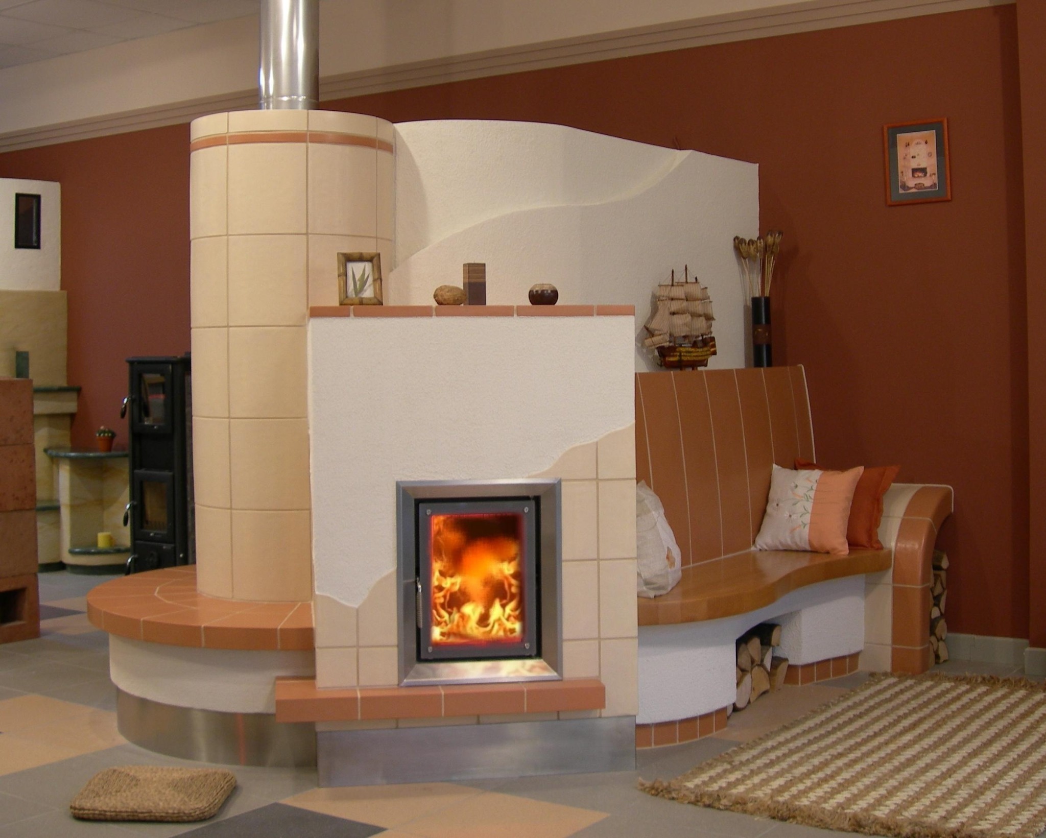 Oryginalny kominek kaflowy z siedziskiem, wykończony w odcieniach beżu i brązu. Ogień płonie za metalowymi drzwiczkami. Dekoracje na półce i dywanik dopełniają wystrój.