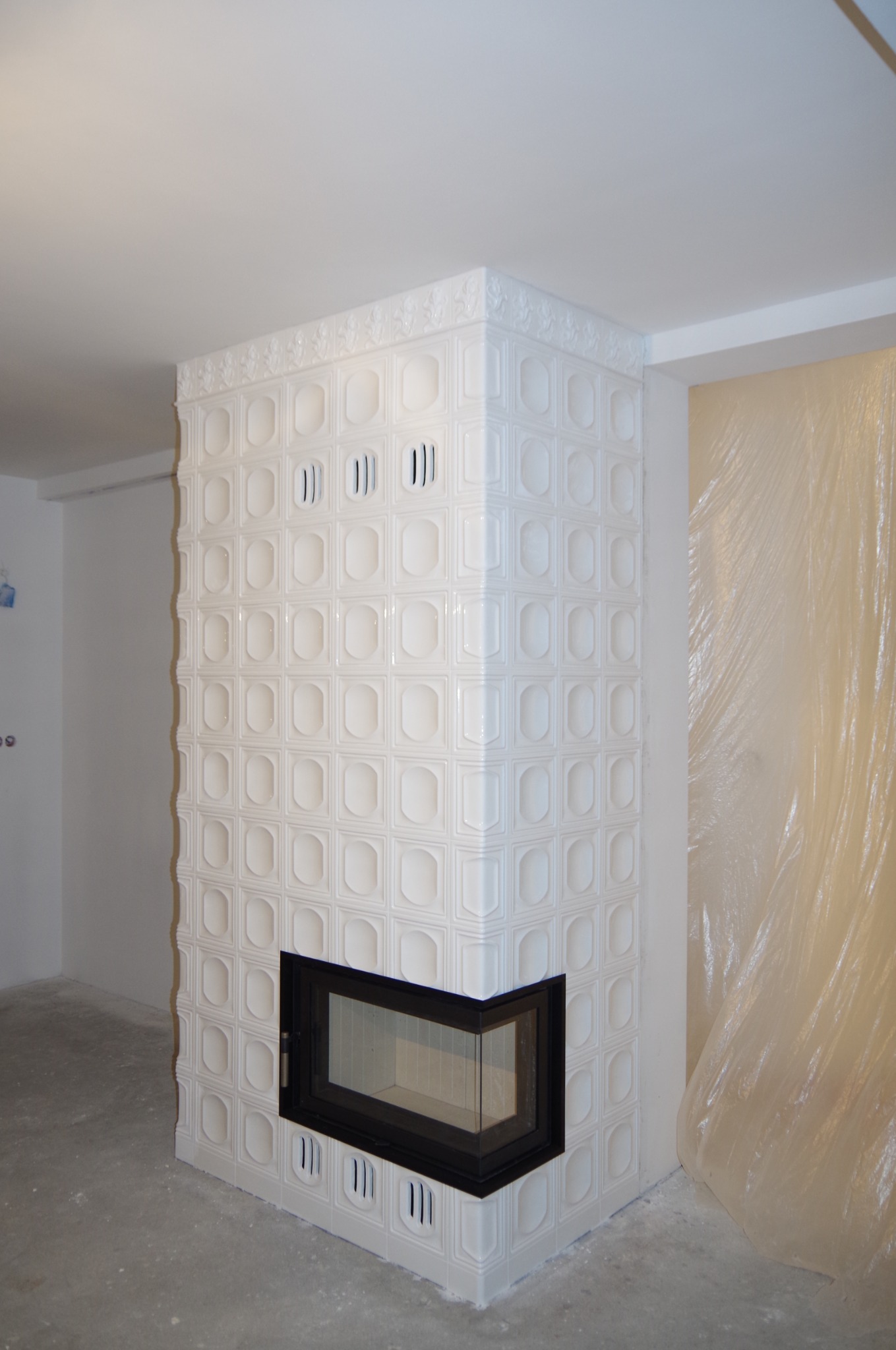 Biały, kaflowy kominek narożny z czarną ramą paleniska, w surowym wnętrzu. Widoczne detale zdobień kafli i kratki wentylacyjne. Część ściany zabezpieczona folią malarską.