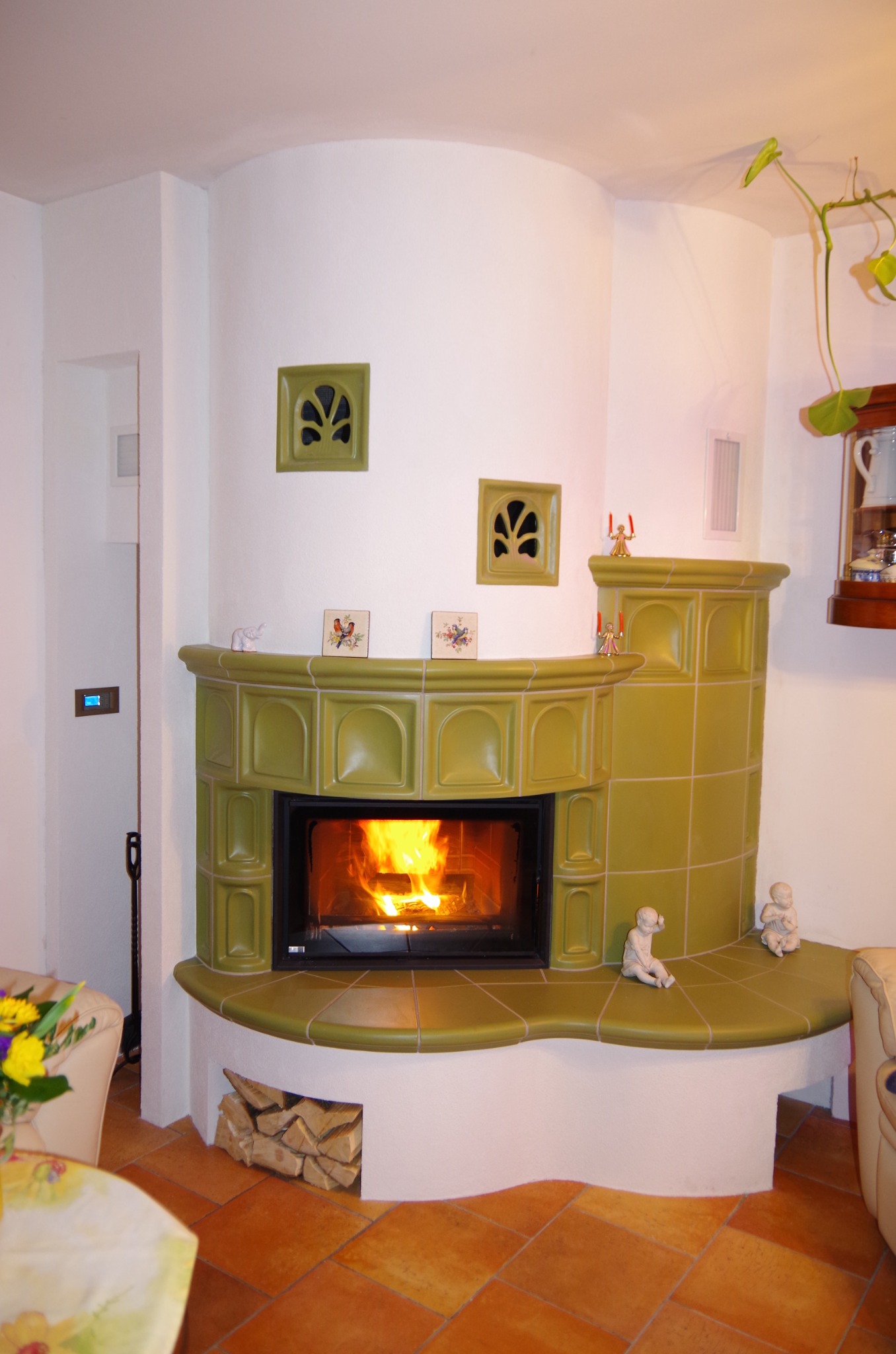 Oliwkowy kominek kaflowy z paleniskiem, ozdobiony figurkami i kaflami z motywem roślinnym, na tle białej ściany i terakotowej podłogi. Widoczne drewno pod paleniskiem.