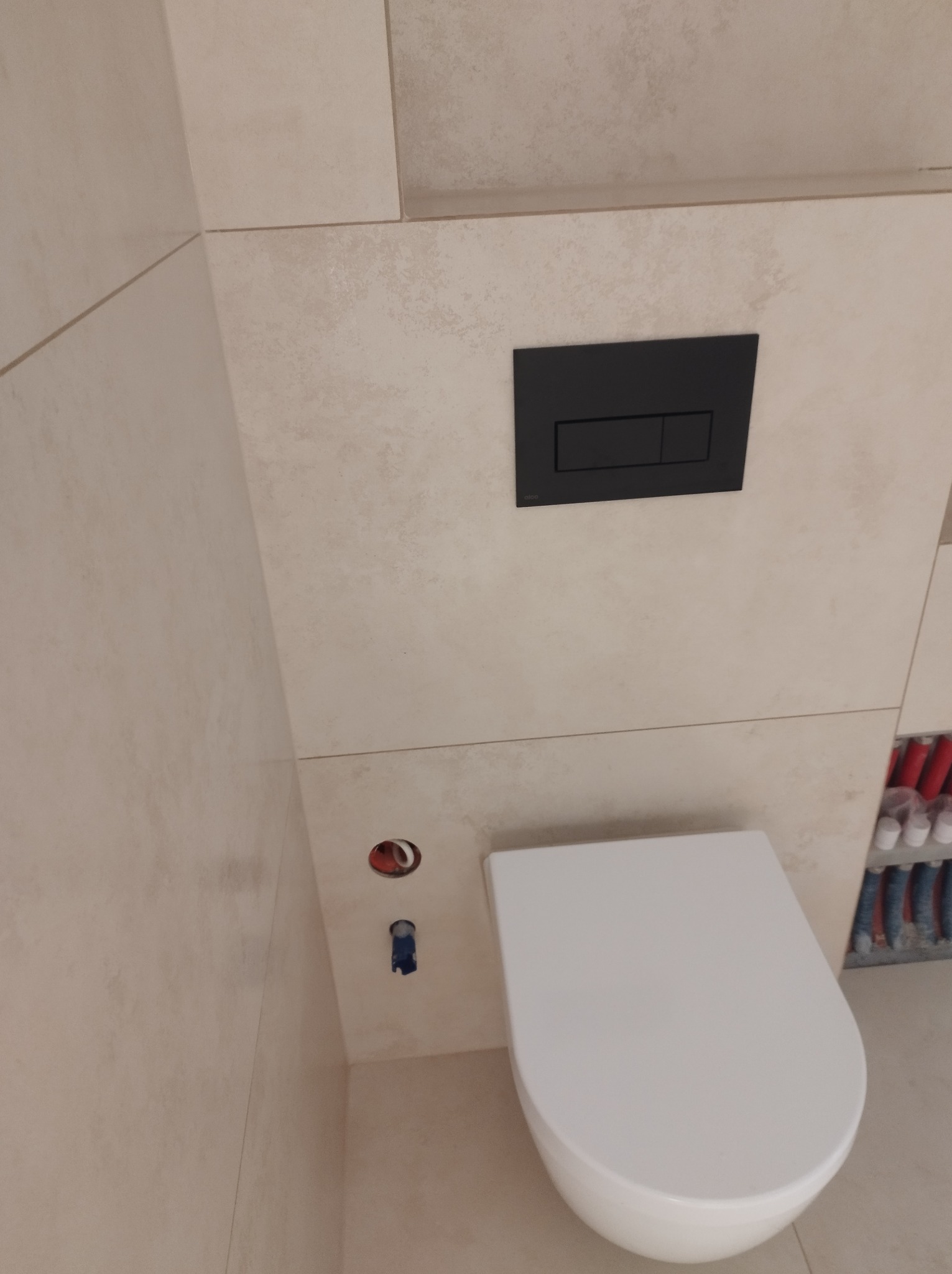 Wisząca toaleta z czarnym przyciskiem spłukującym na beżowych płytkach. Widoczne przyłącza wody i kanalizacji. Minimalistyczny design łazienki po remoncie.